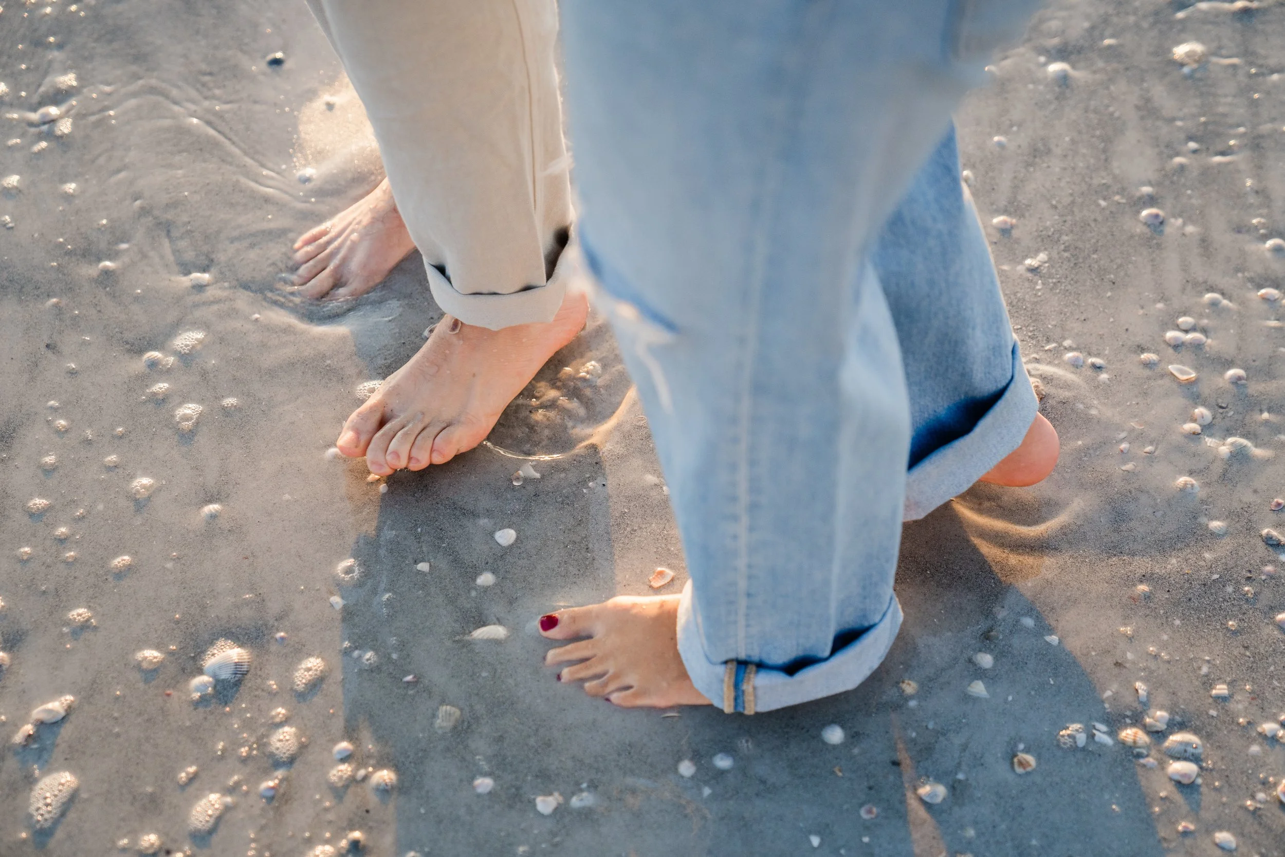 Person standing barefoot on sandy beach with small seashells, wearing rolled-up light blue jeans and a beige jacket.