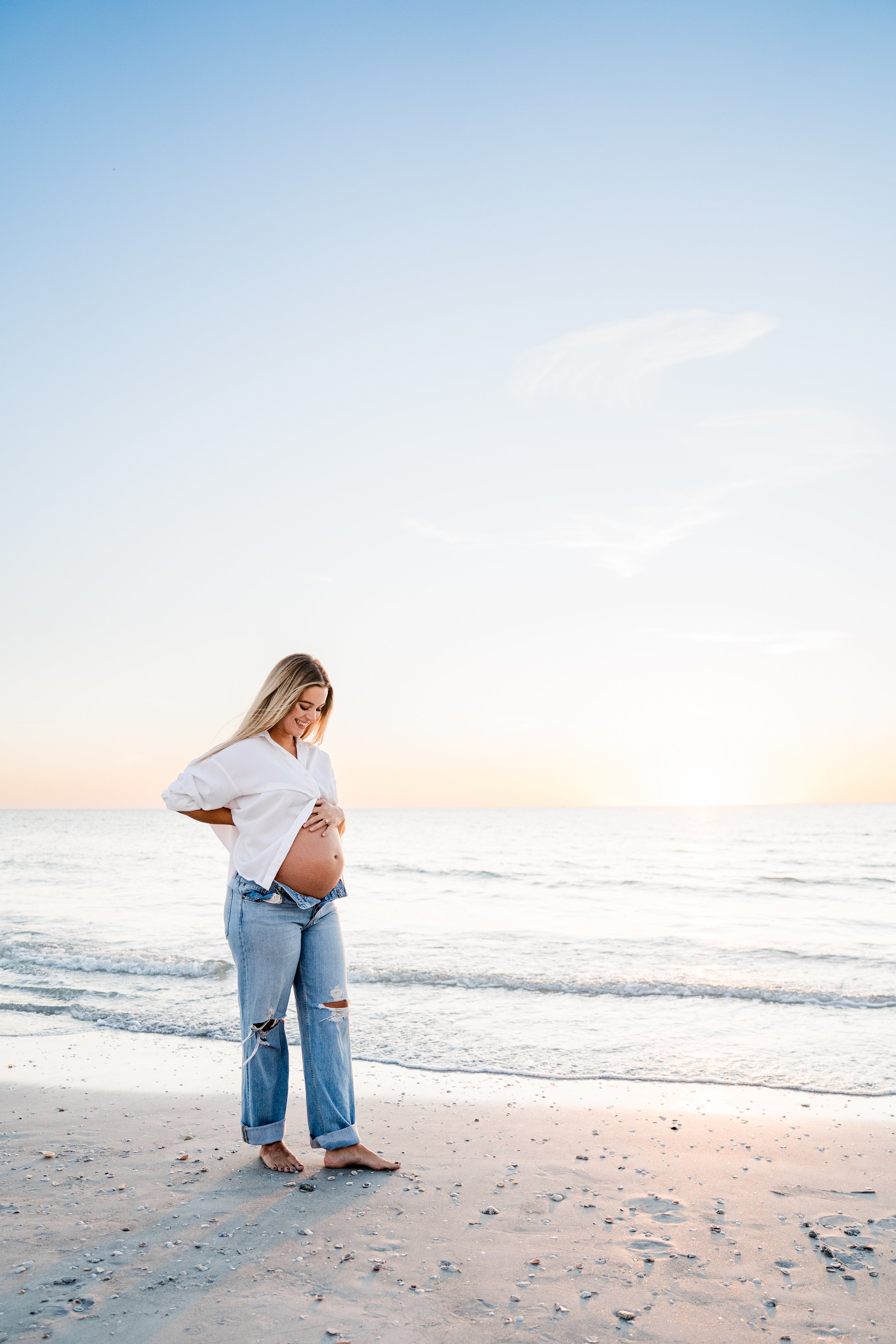 A pregnant woman stands barefoot on the beach at sunset, smiling and looking down at her belly, wearing ripped jeans and a white shirt.