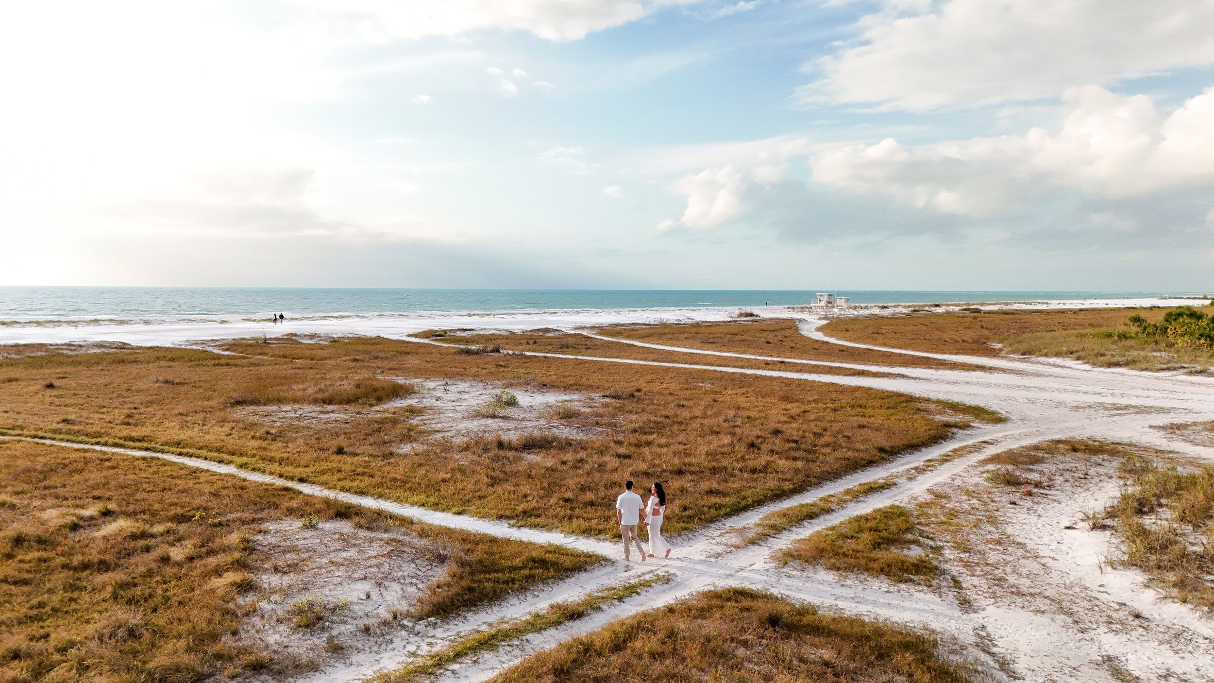 A couple walking hand in hand on a sandy path through grassy dunes towards the beach with the ocean in the background. The sky is partly cloudy.