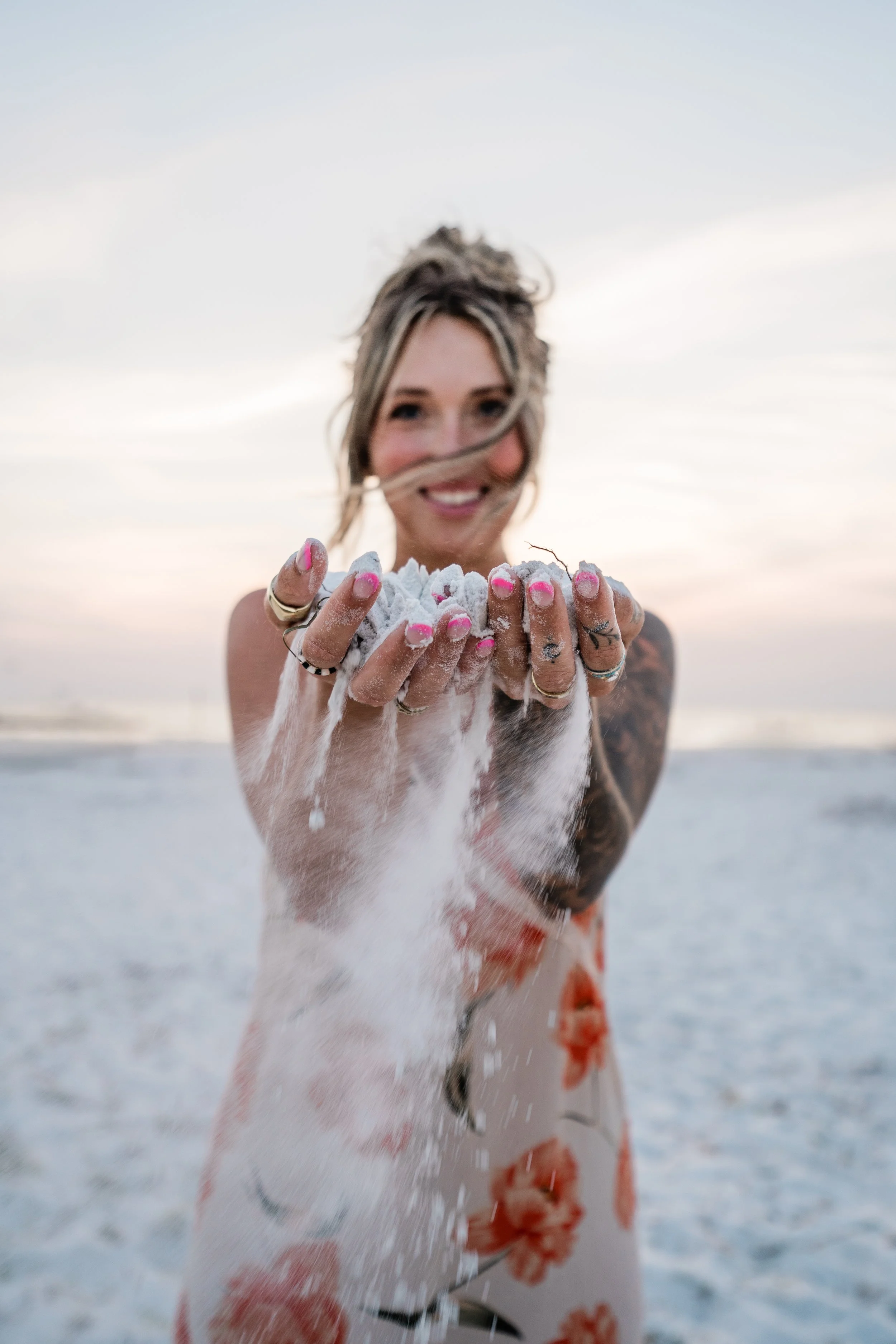 Woman on the beach holding sand in her hands, smiling at the camera, wearing a floral dress.