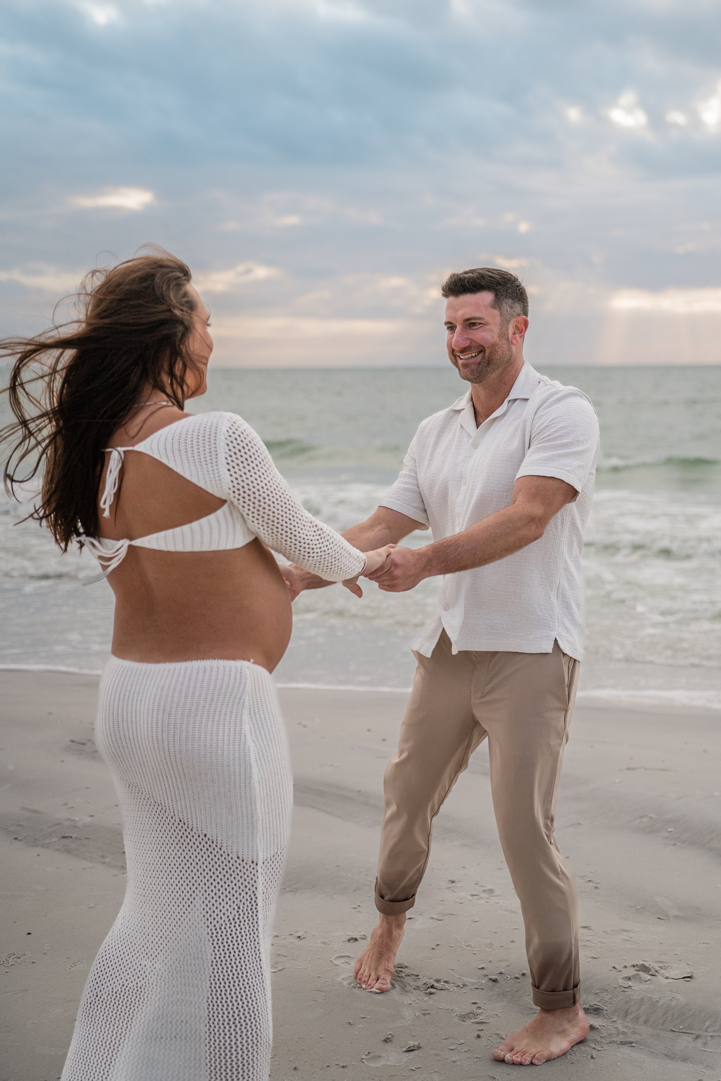 A pregnant woman and a man holding hands on the beach at sunset, smiling and facing each other.