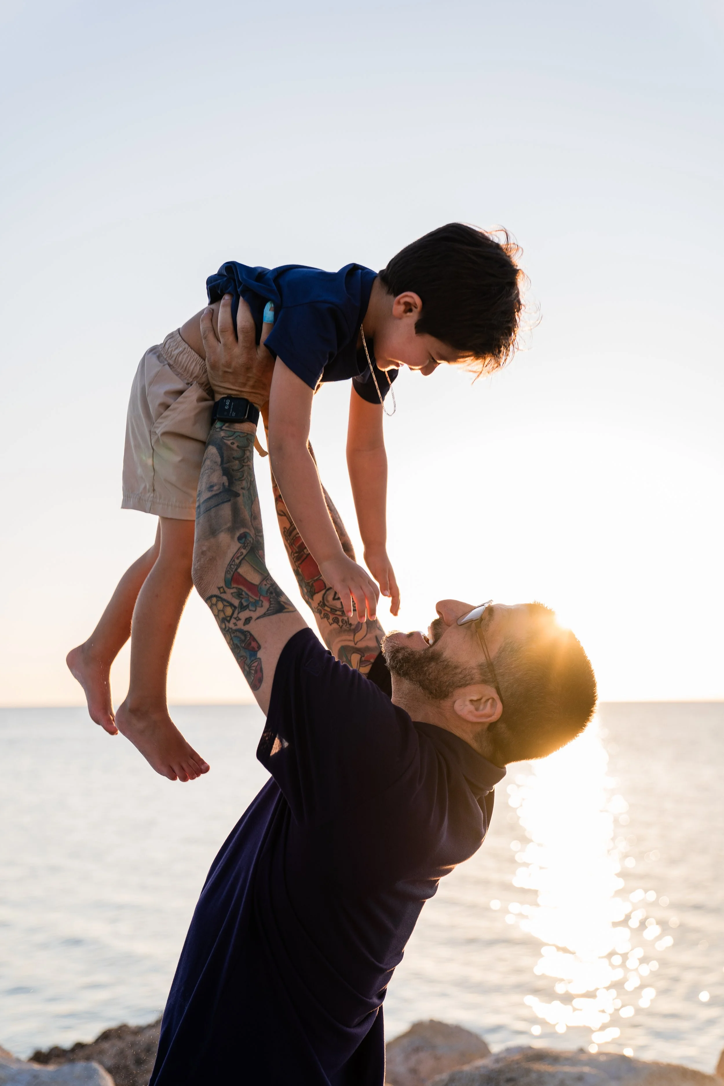 A man with tattoos lifting a young boy with dark hair, wearing a navy shirt and beige shorts, above his head by the water during sunset.