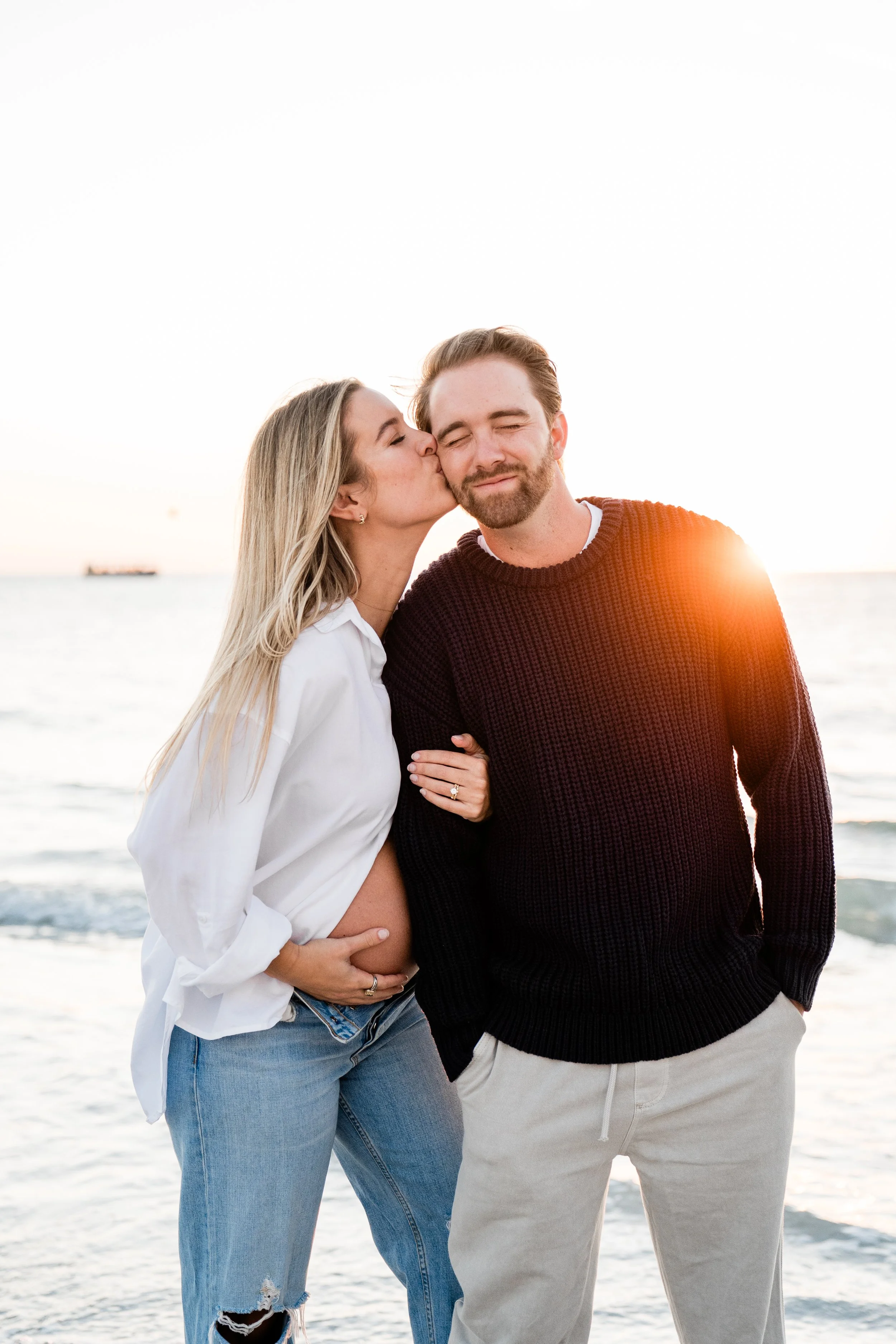 A pregnant woman kissing a man on the cheek at the beach during sunset