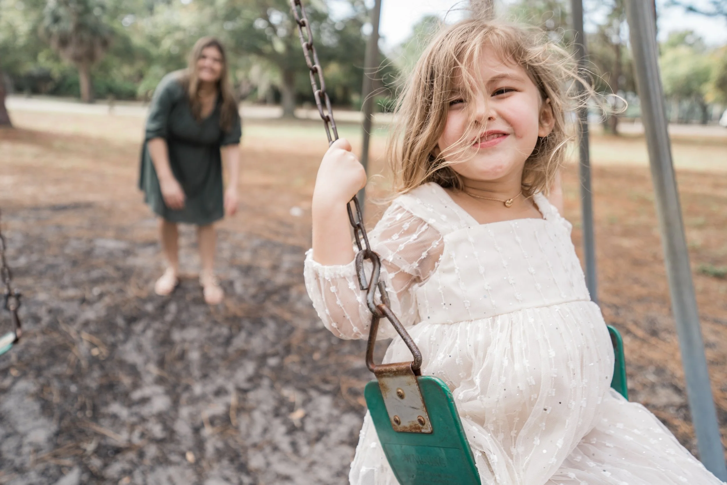 Young girl smiling on a green swing, with woman in background in a park with trees.