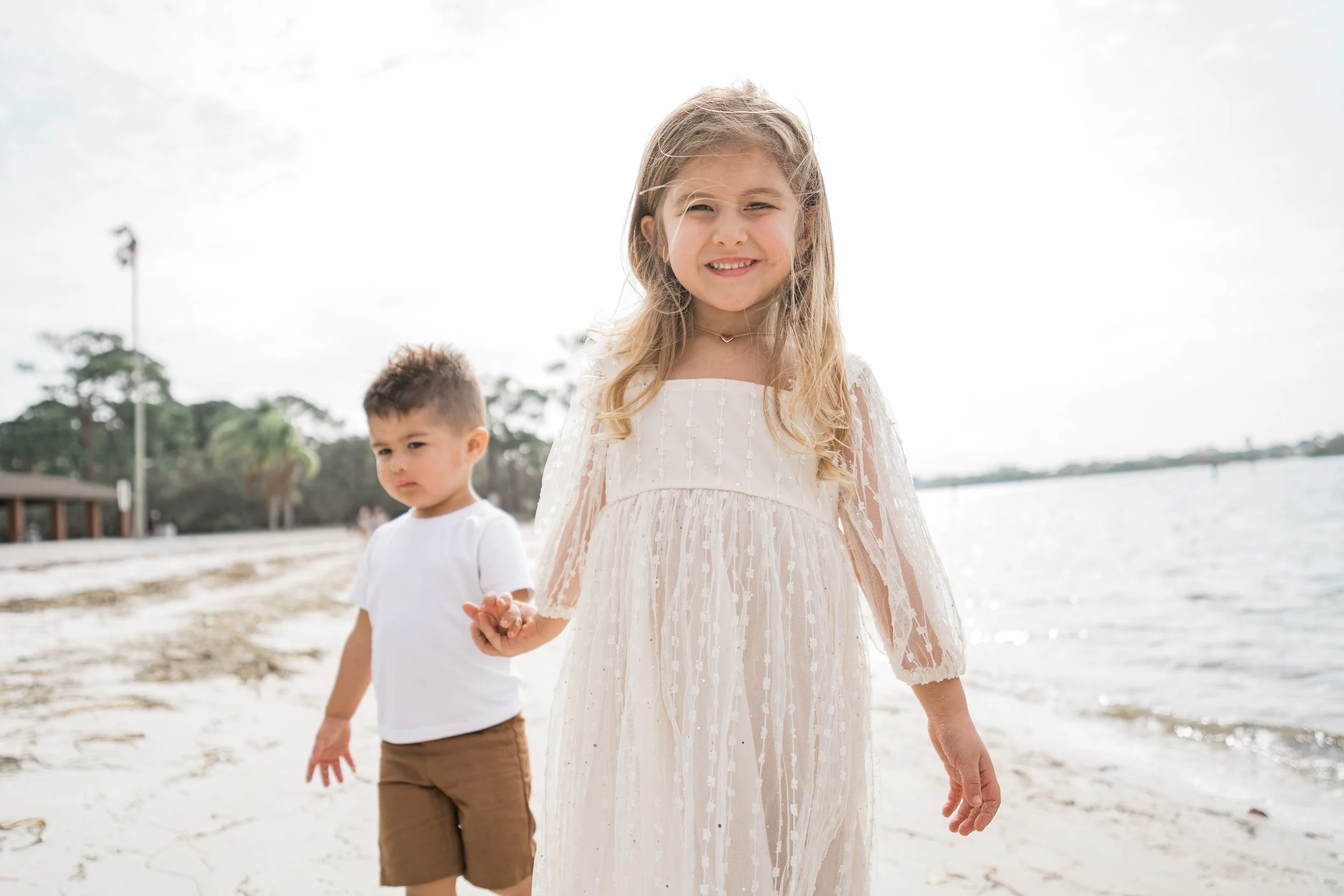 A young girl in a white dress holding hands with a young boy in a white t-shirt and brown shorts, walking on a beach.