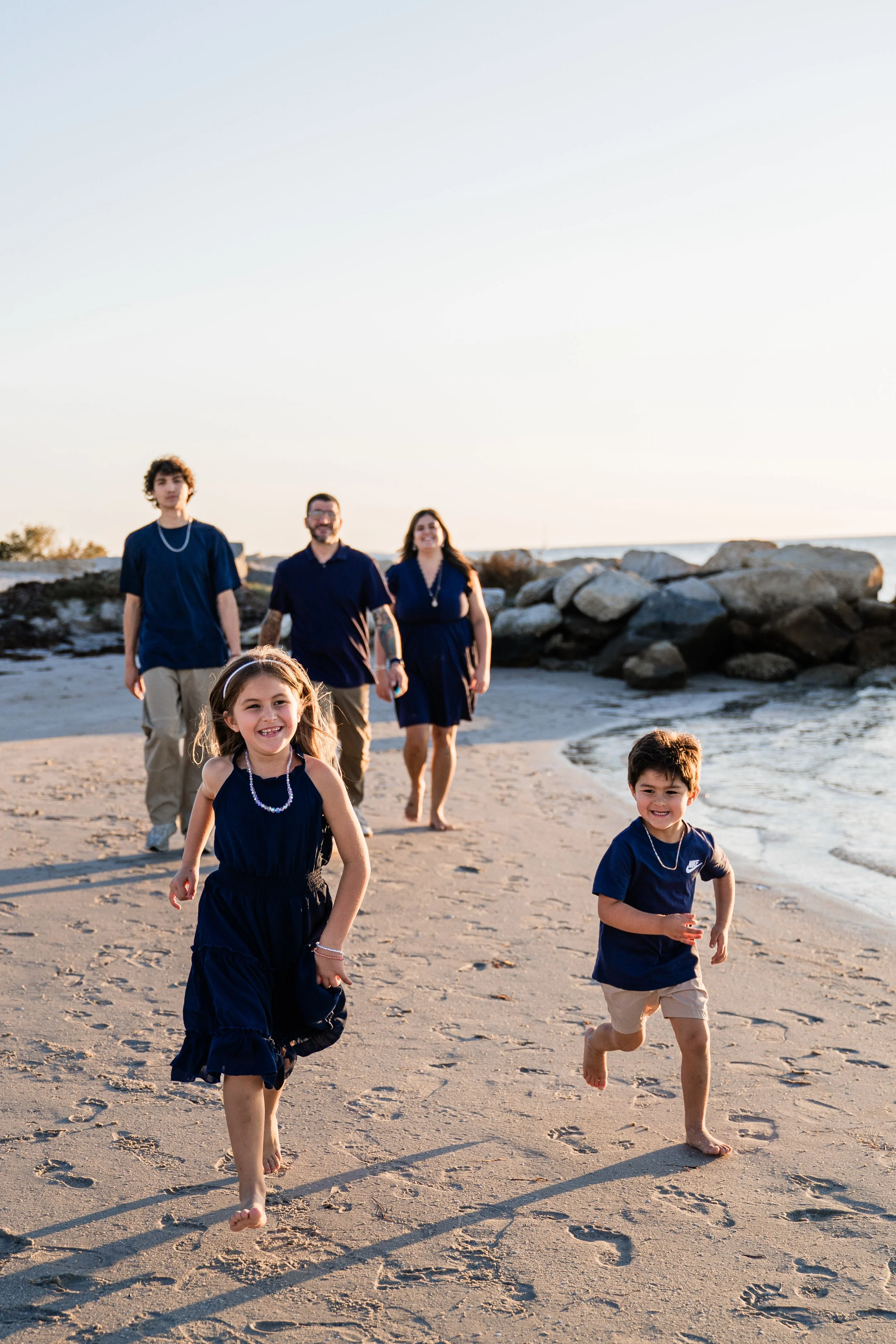 A family of six walking and running on a sandy beach near rocks, dressed in matching navy blue outfits, during sunset.