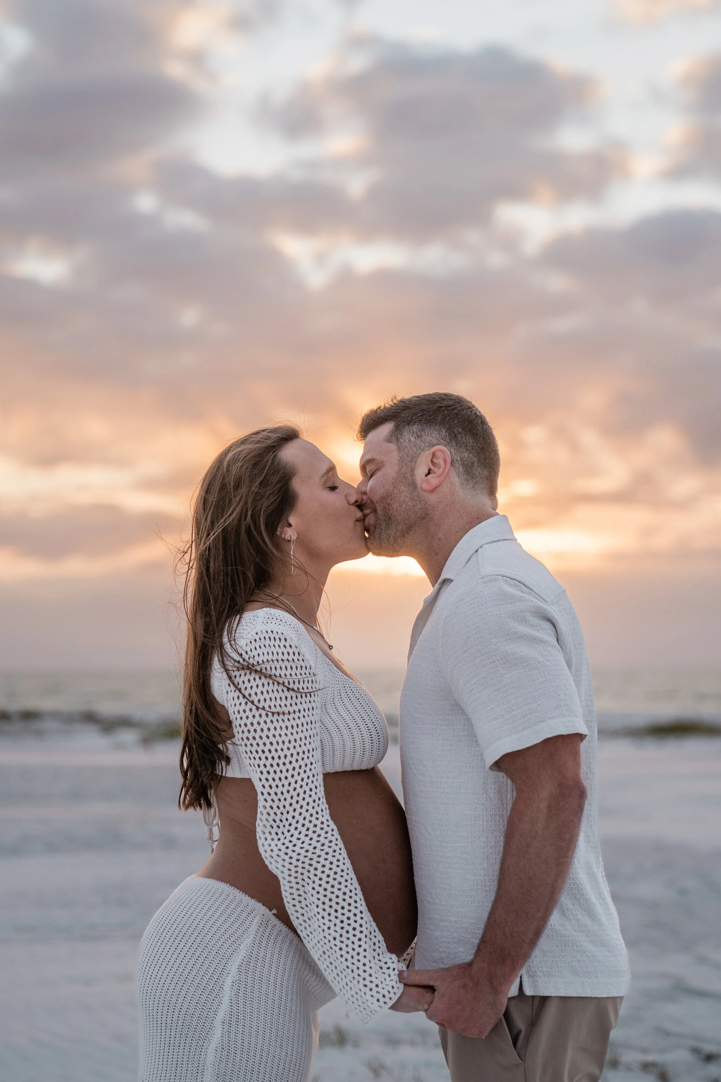 A couple kissing on the beach at sunset, with the woman being pregnant, holding hands, both dressed in white.