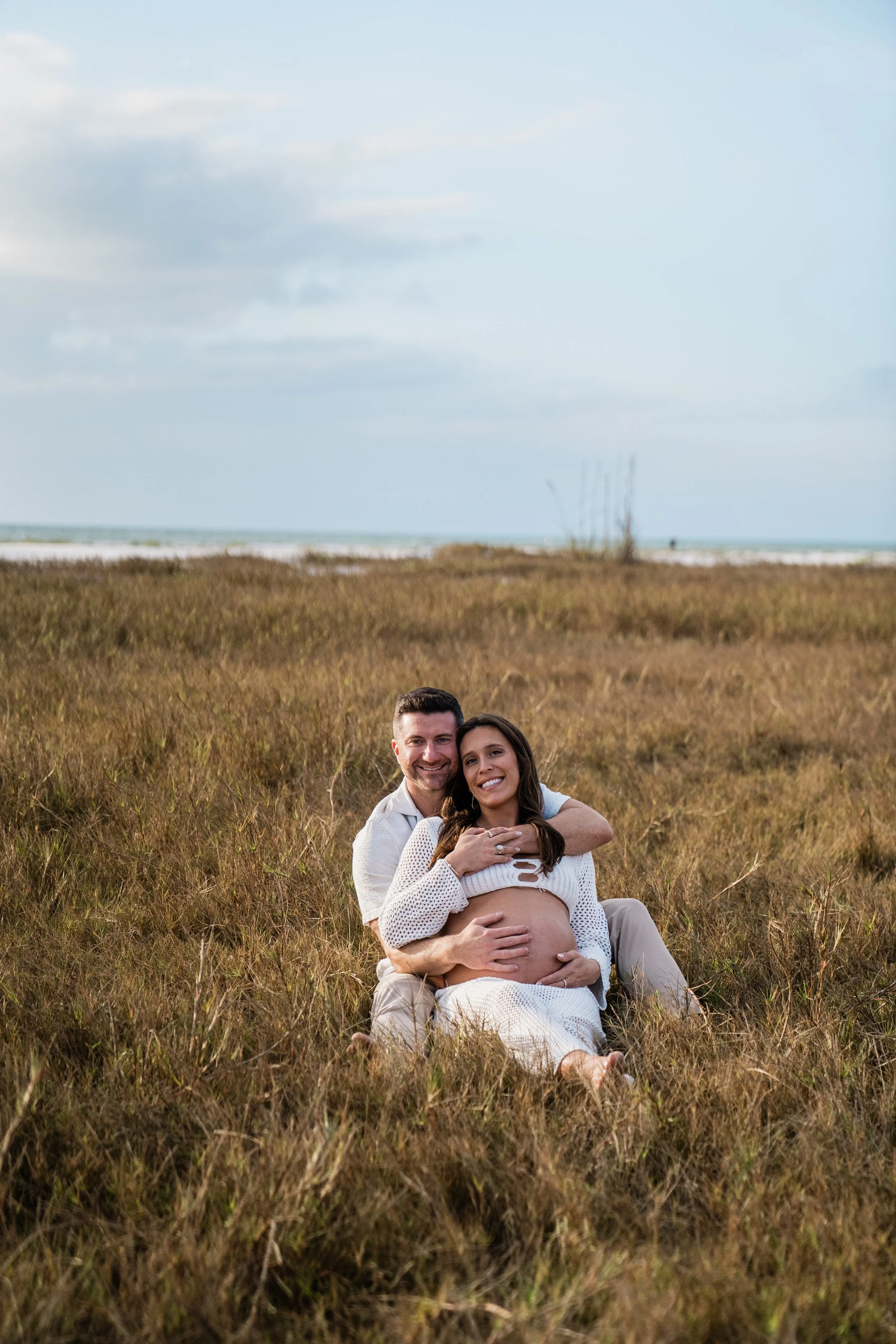 A happy couple is sitting on the grass at the beach, with the woman showing her pregnant belly, both smiling at the camera.