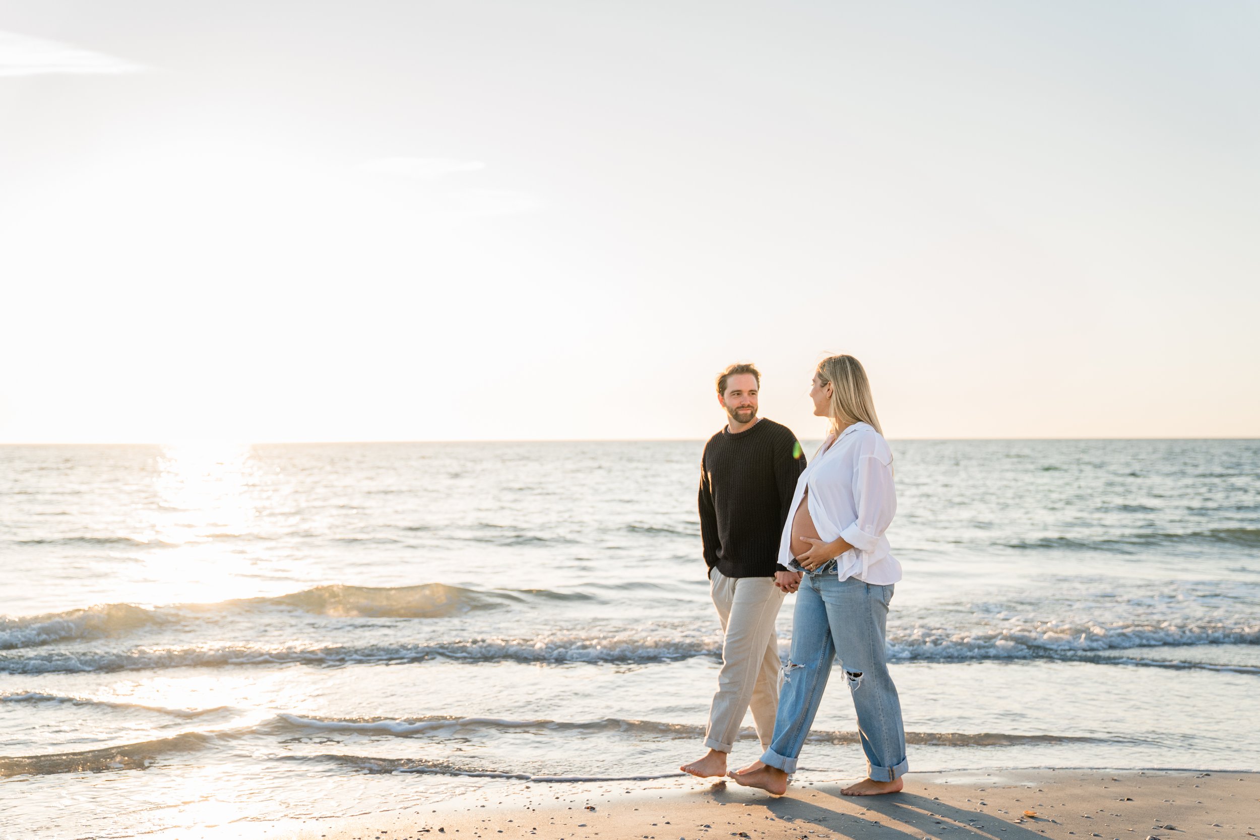 A pregnant woman and a man walking barefoot on the beach during sunset, holding hands and looking at each other.