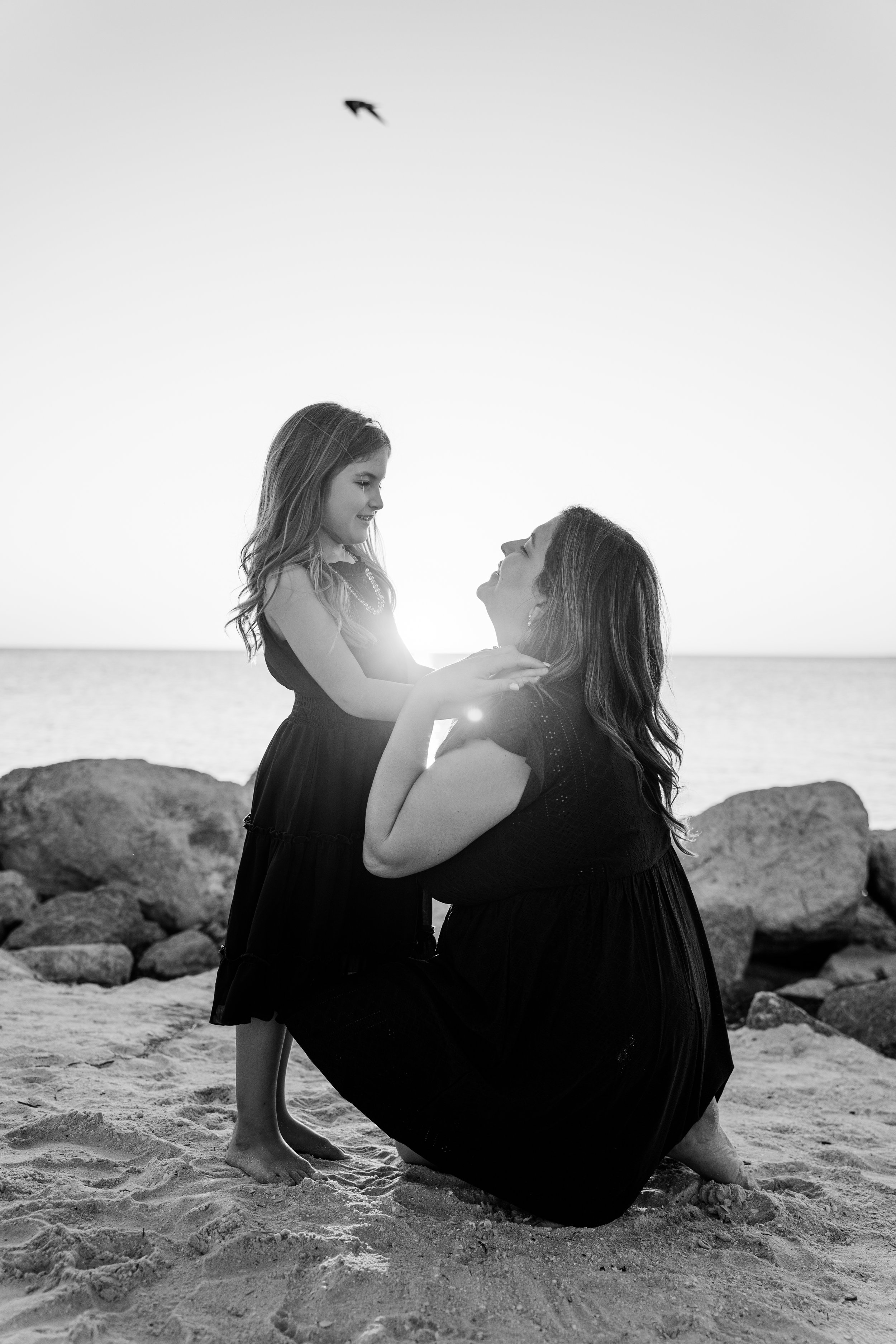 A woman kneeling on the sand at the beach holding a young girl, both smiling at each other, as the sun sets in the background and a bird flies overhead.