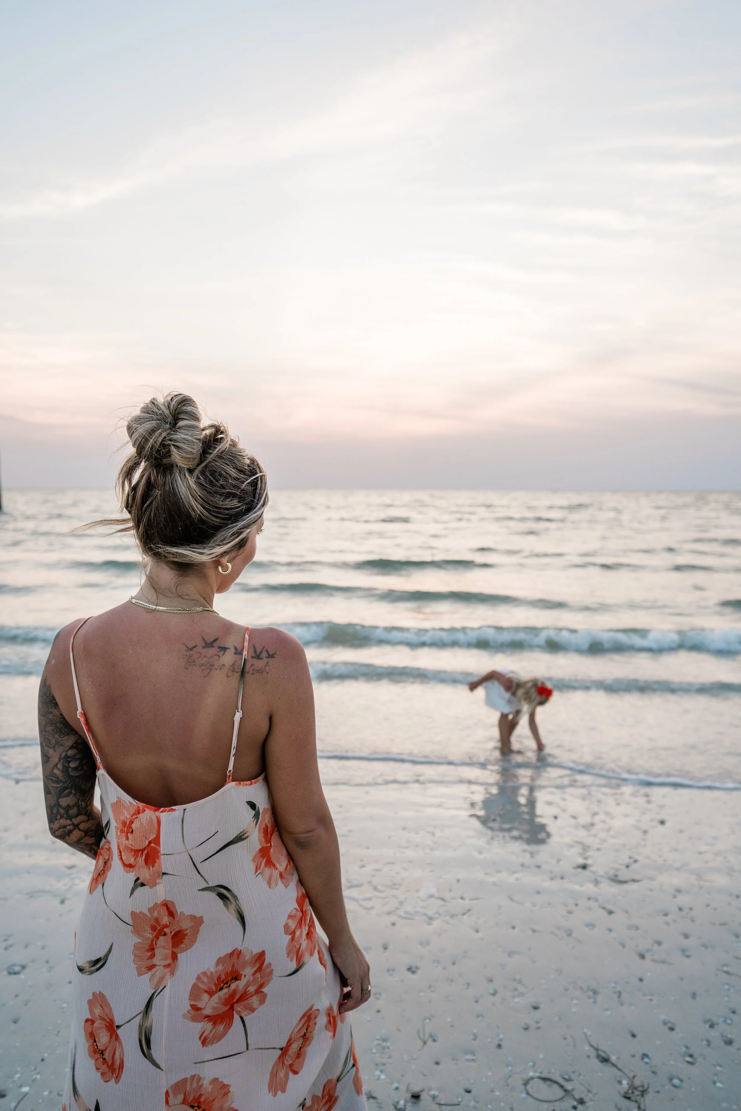 A woman with tattoos on her back and left arm, wearing a white dress with orange flowers, stands on the beach and looks at a person in white clothes bending down in the shallow ocean water with a sunset sky in the background.