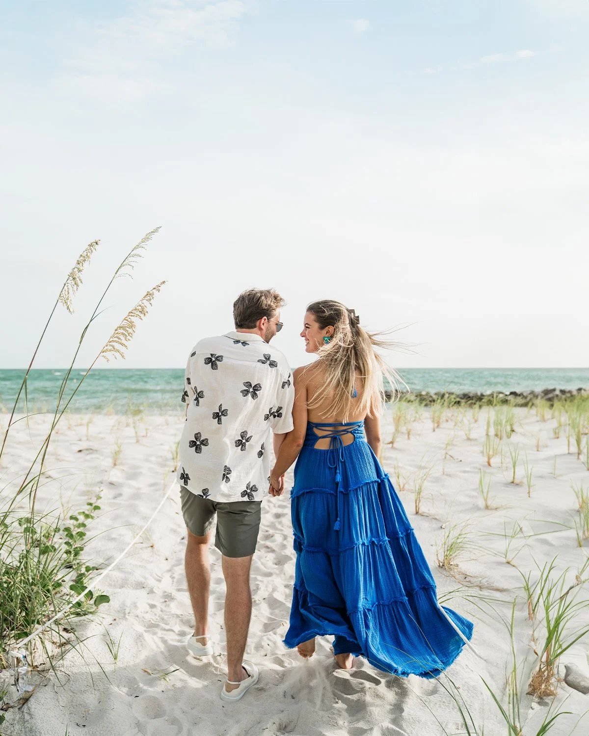 A couple walking hand in hand on a sandy beach with ocean in the background, wearing summer clothes and smiling.