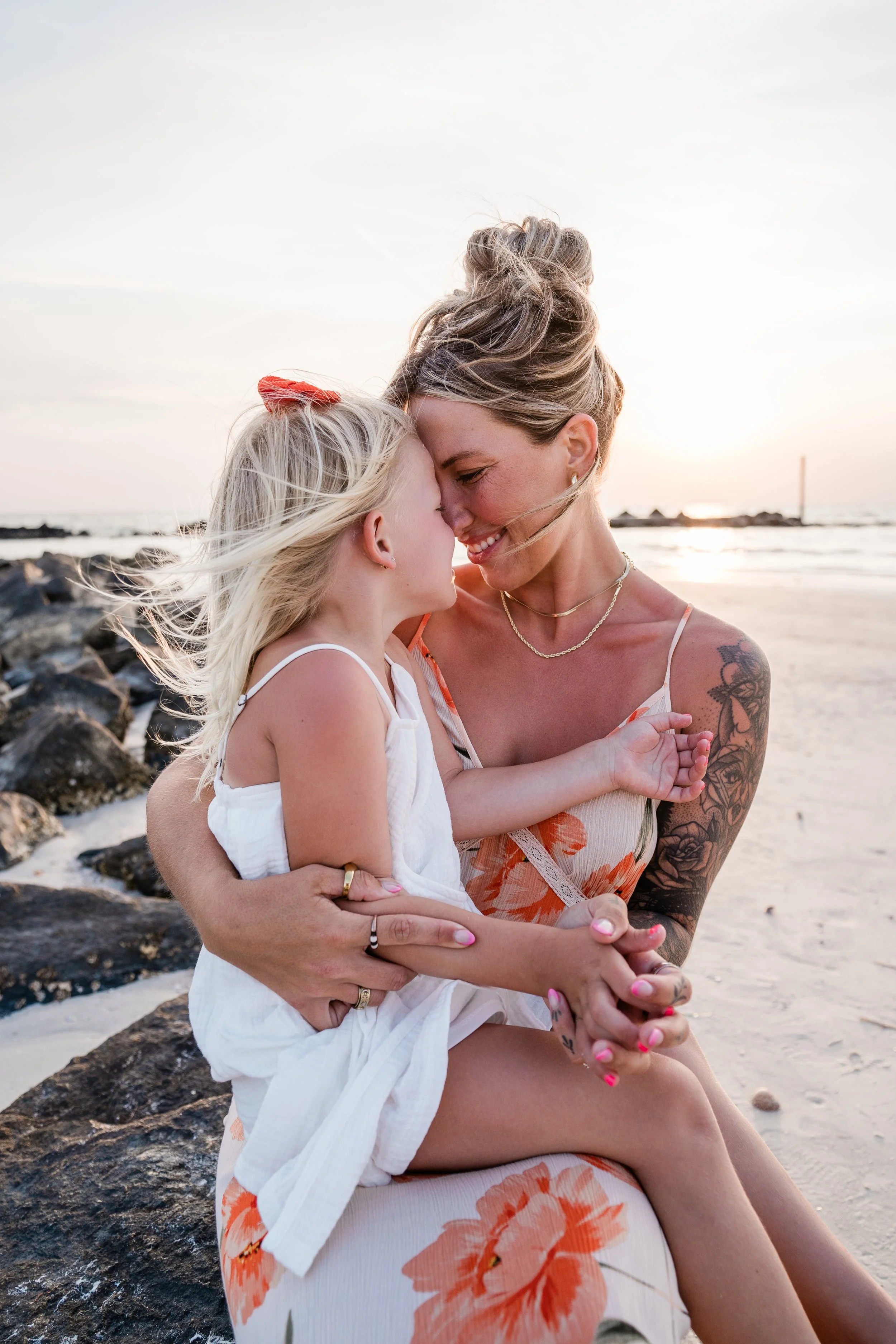 A woman and a girl sit close together on a beach at sunset, smiling and touching foreheads.
