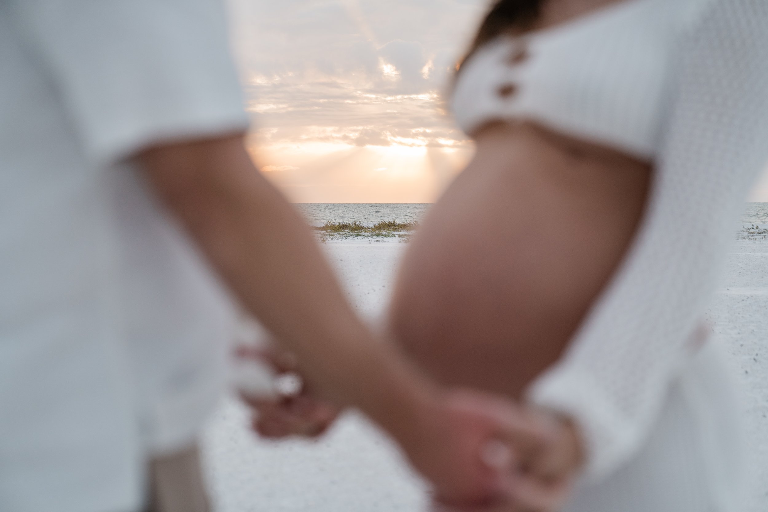 People forming a heart shape with their hands on a beach during sunset, with the ocean and sky in the background.