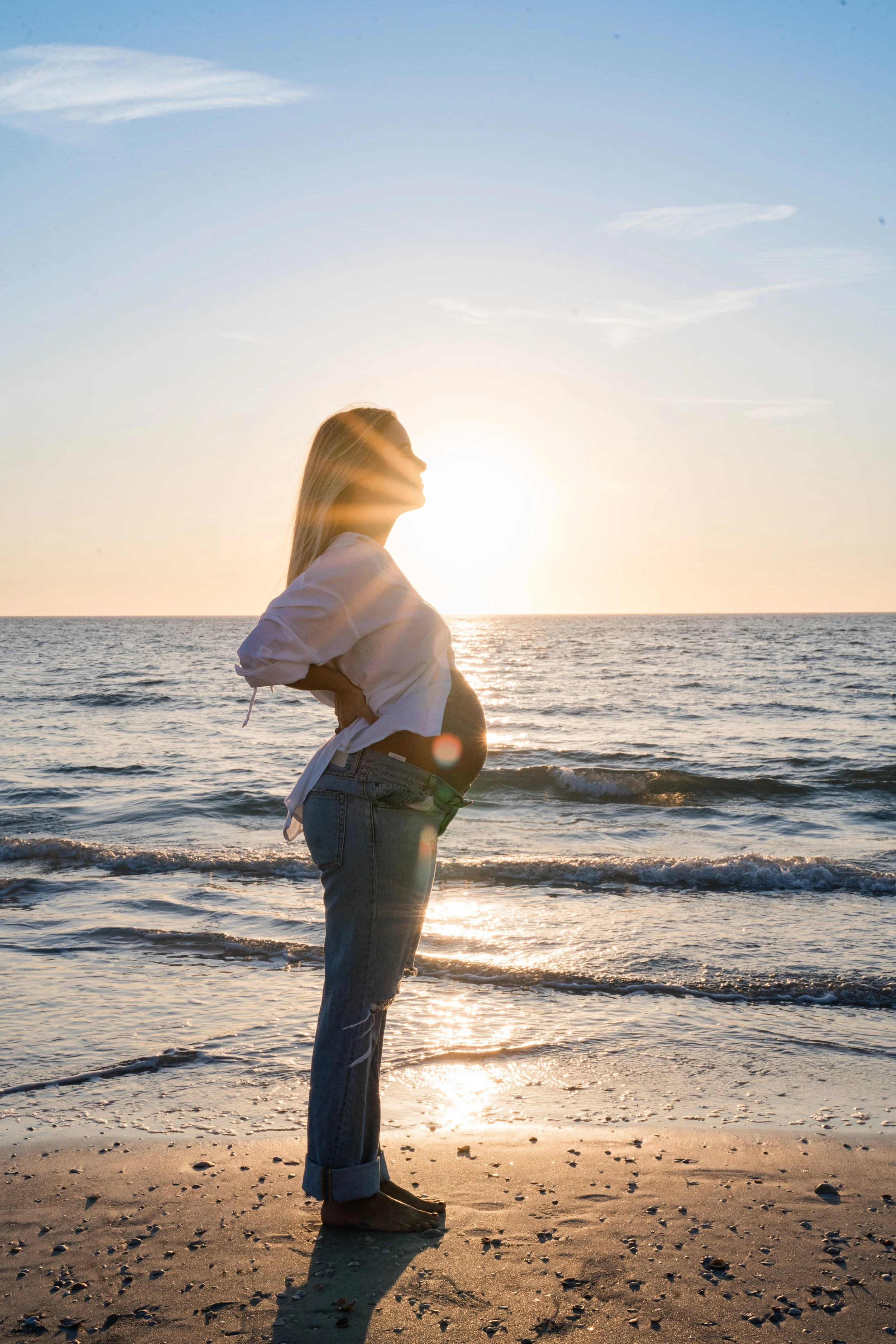 A pregnant woman stands on the beach at sunset, facing the ocean with her hands on her hips, wearing jeans and a white shirt.