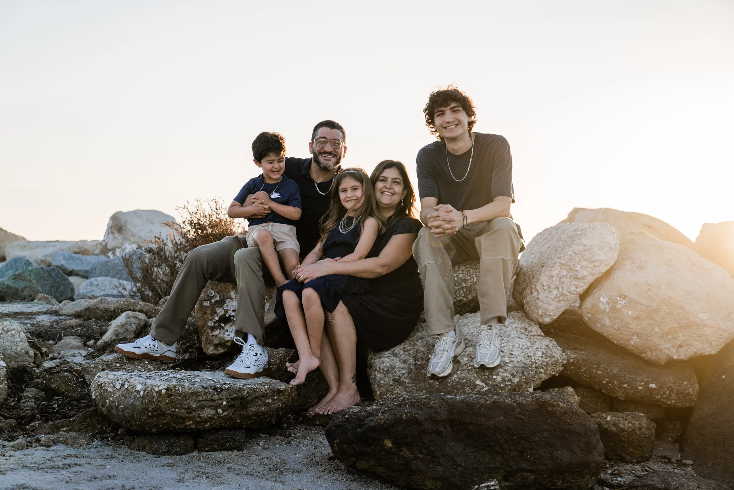 Family of five sitting on rocks outdoors during sunset, smiling at the camera.