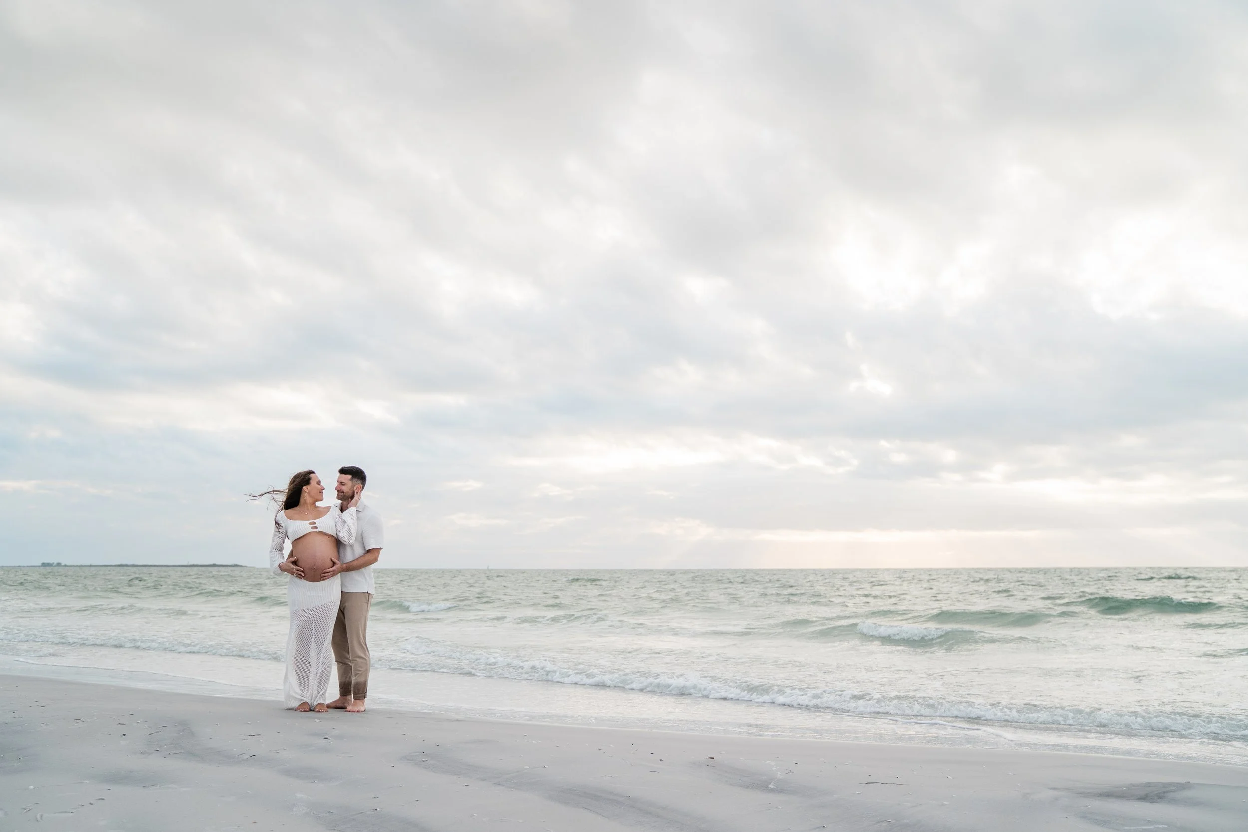 A pregnant woman and a man standing on the beach, holding her belly and looking at each other, with the ocean and cloudy sky in the background.