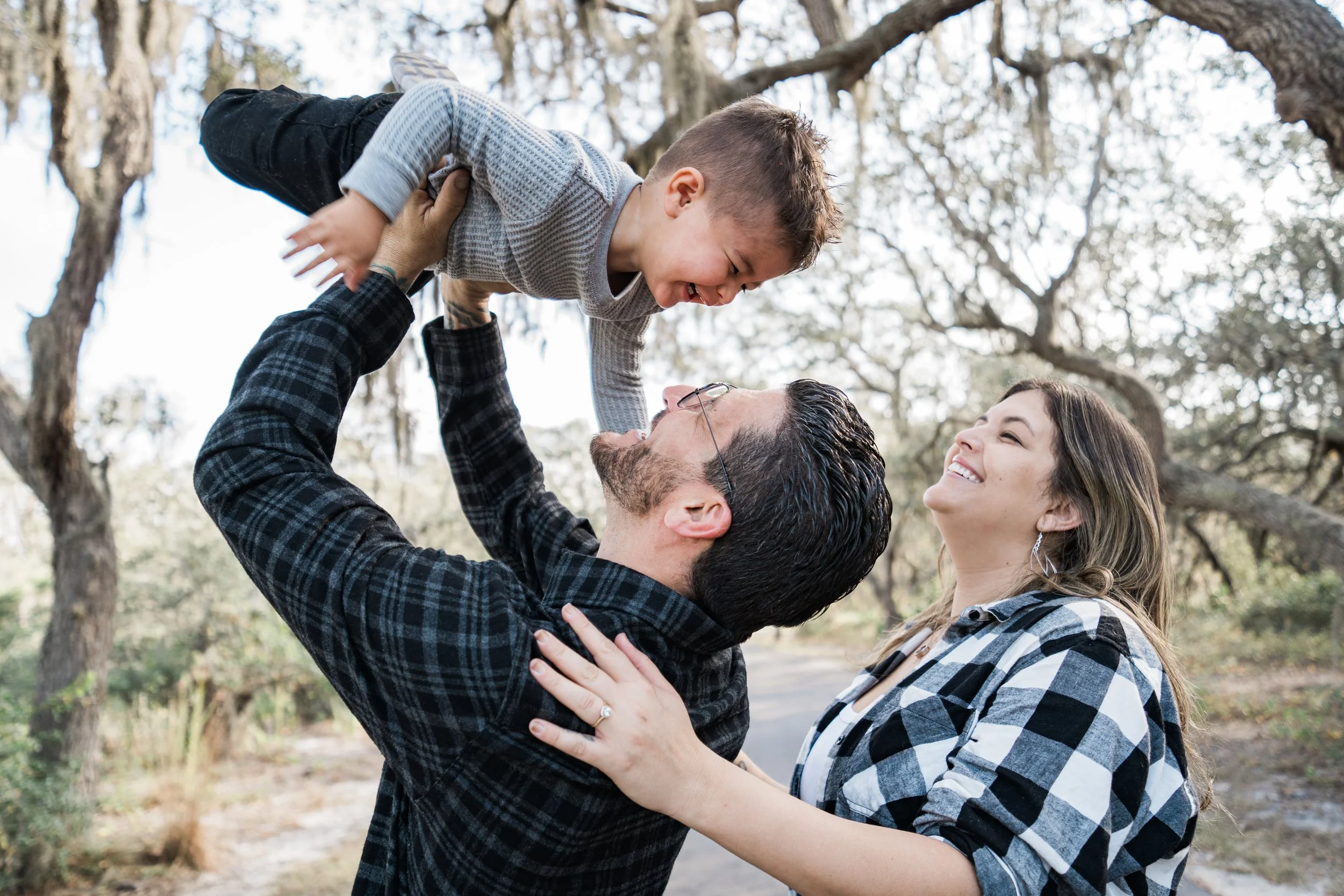 A family of three outdoors in a wooded area, with the father lifting and playing with his son while the mother looks on smiling.