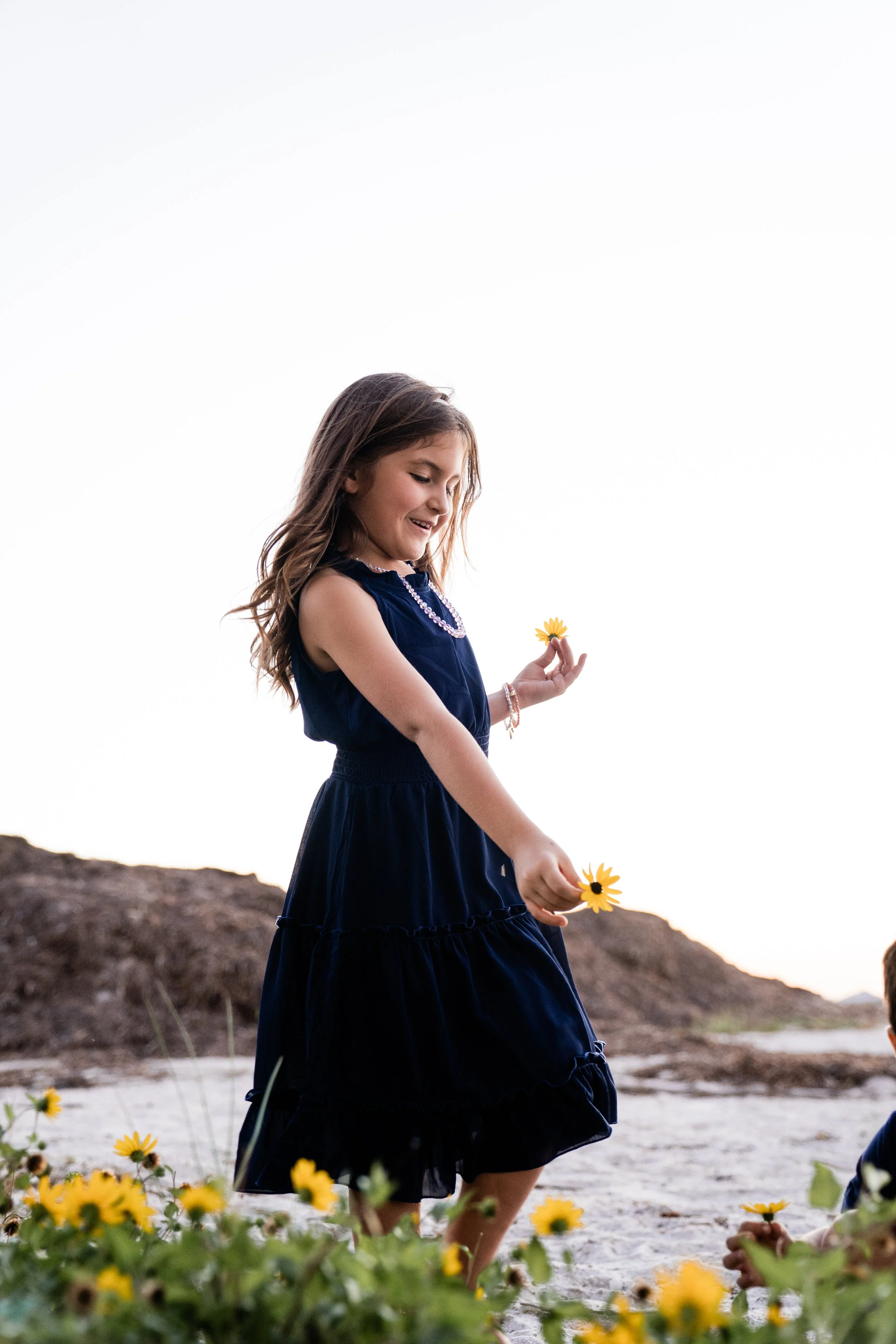 A young girl in a navy dress holding yellow flowers and standing outdoors near rocks, smiling.