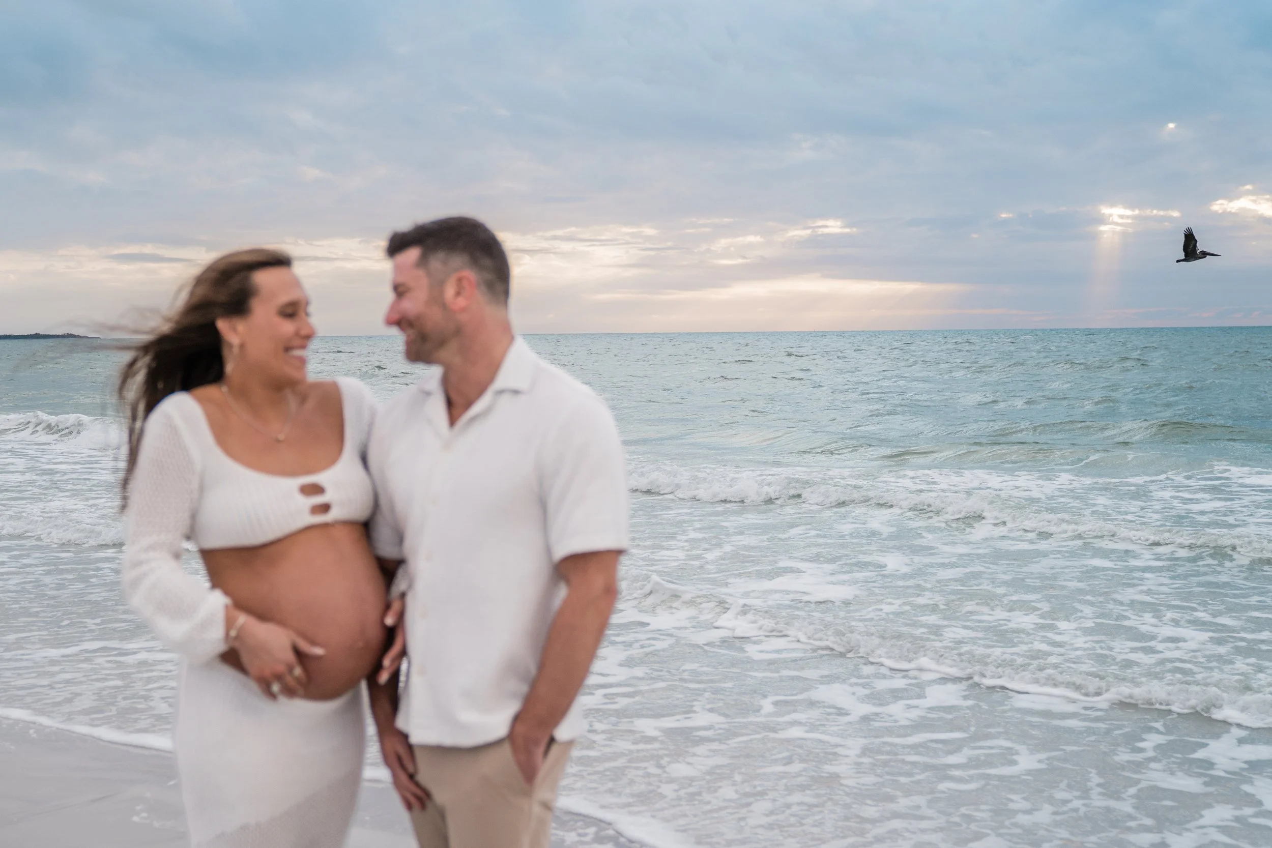 A pregnant woman and a man stand on a beach near the ocean, smiling and looking at each other, with the sky and sea in the background.