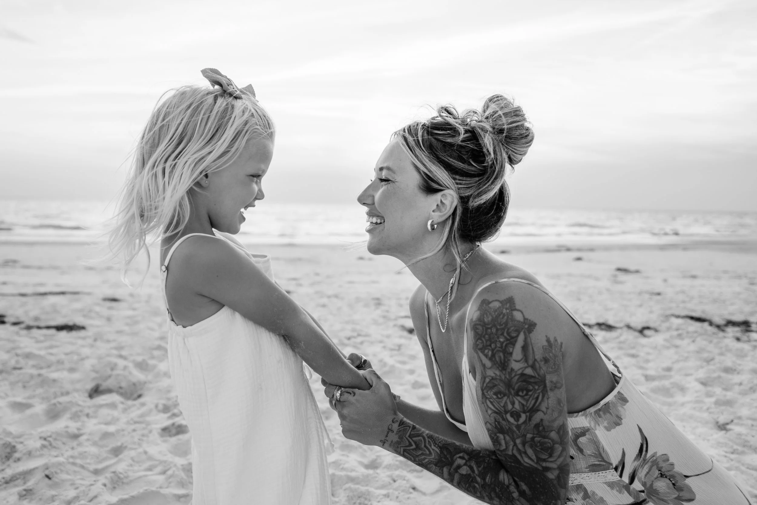 A woman with tattoos and a bun hairstyle holding hands with a young girl on a beach, both smiling and looking at each other.