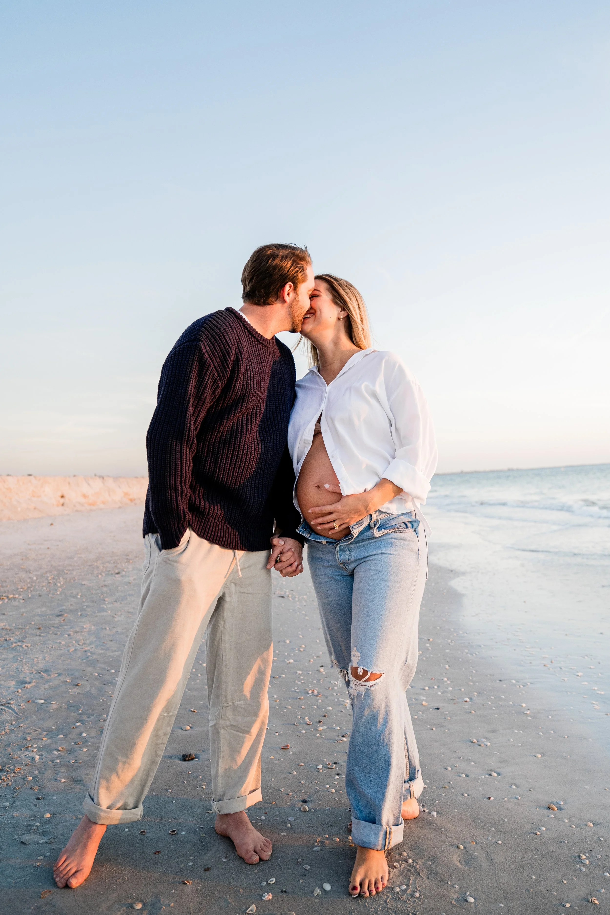 A pregnant woman and her partner are kissing on the beach during sunset.
