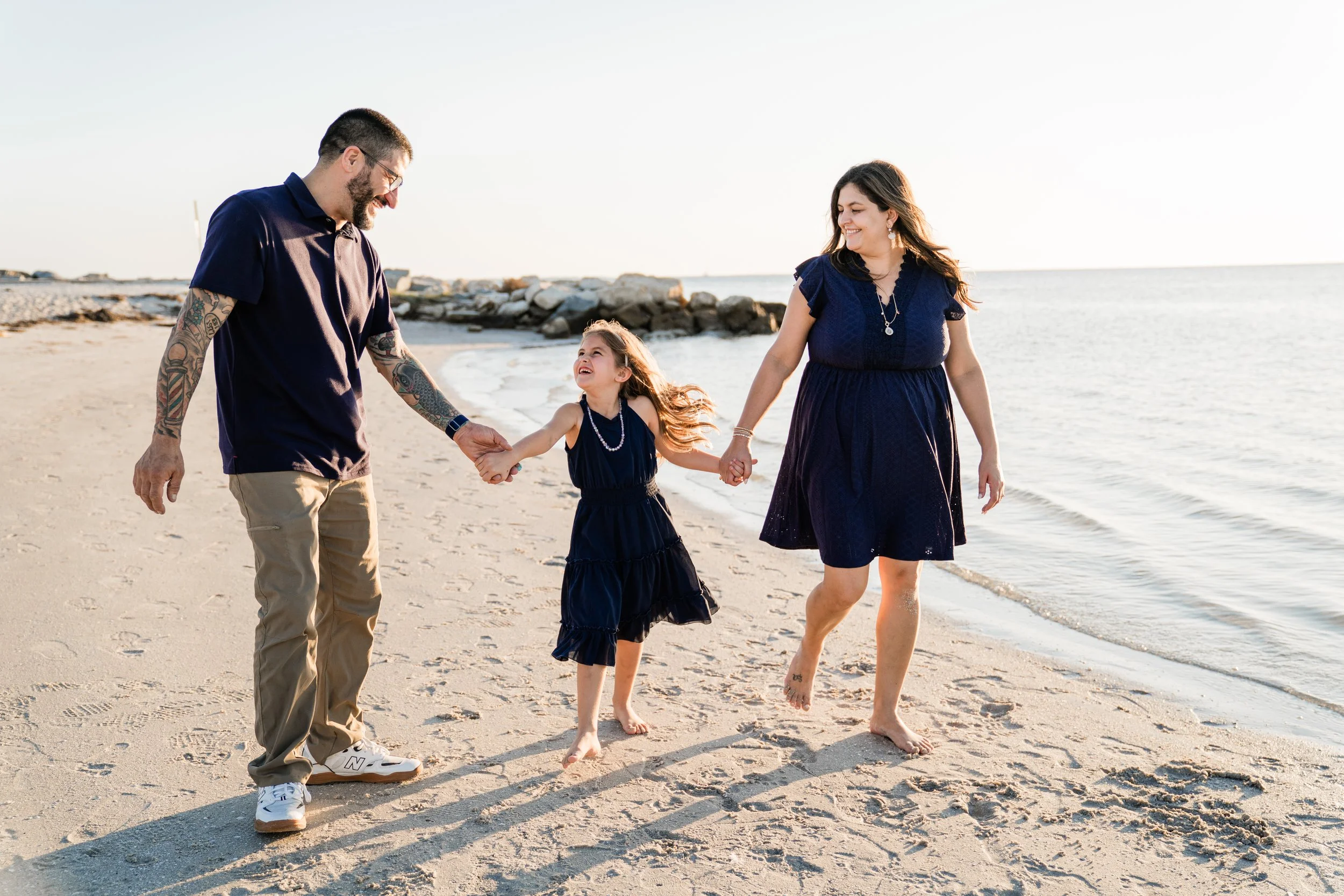 A family of three walking on a beach, holding hands and smiling, during sunset.