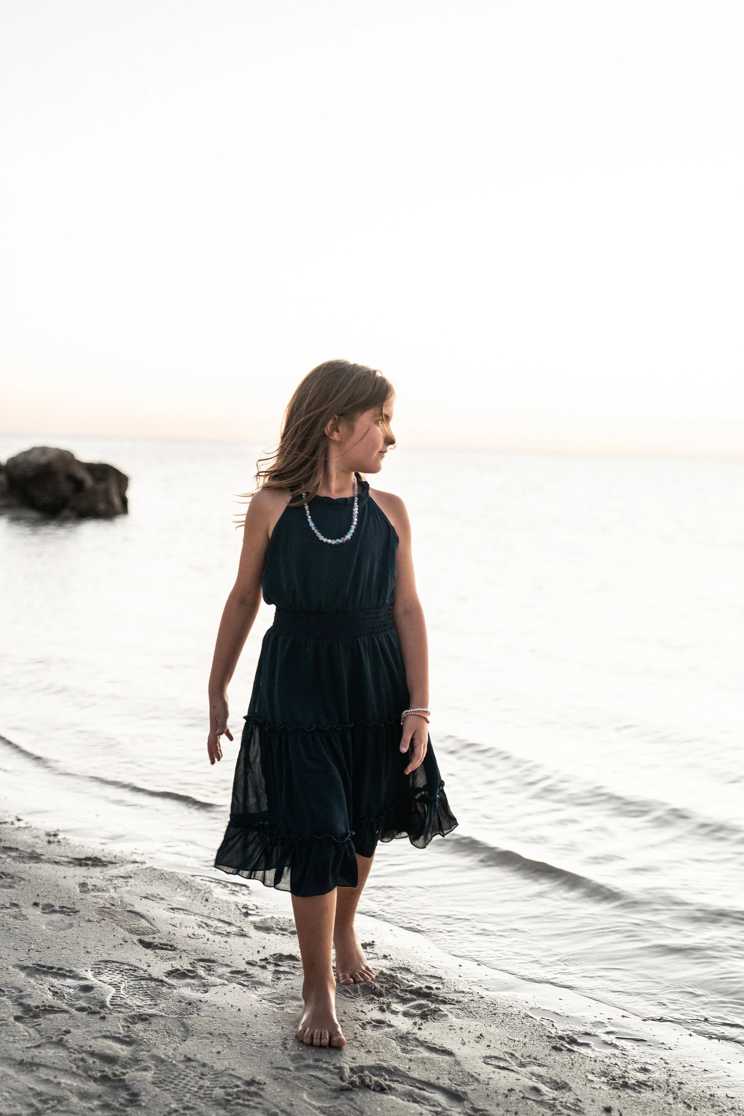 Young girl in a black dress walking barefoot on the beach at sunset.