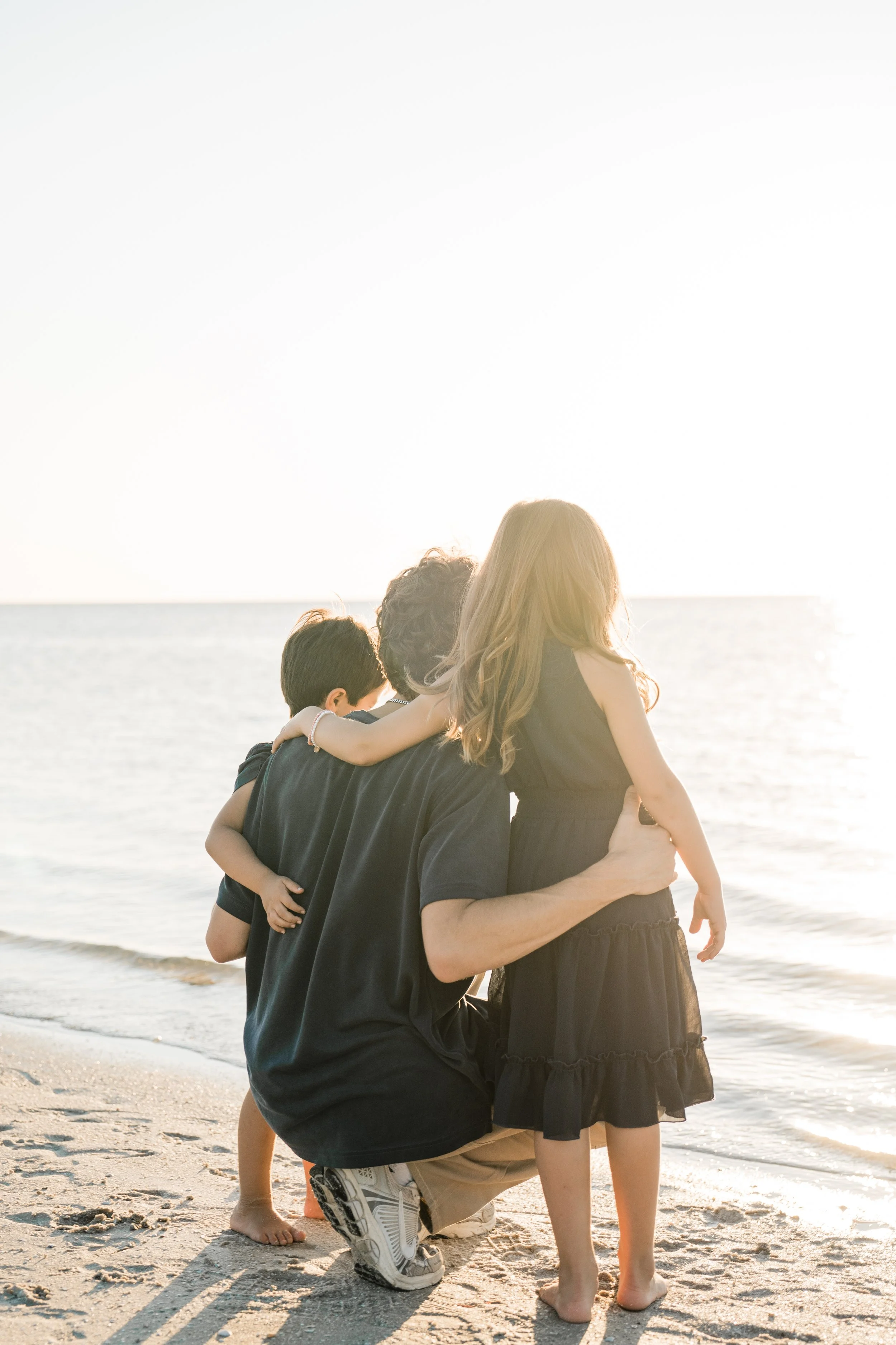 A family of three at the beach during sunset, with a man kneeling and holding a young girl, and another child standing beside them, all facing the ocean.