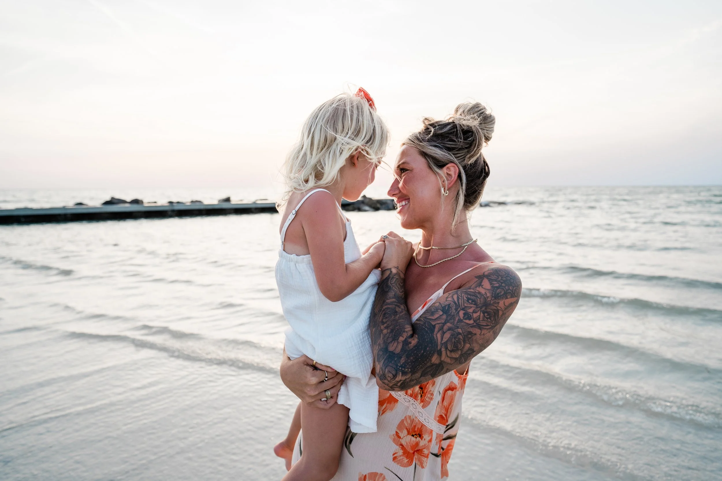 A woman with tattoos holding a young girl at the beach during sunset.