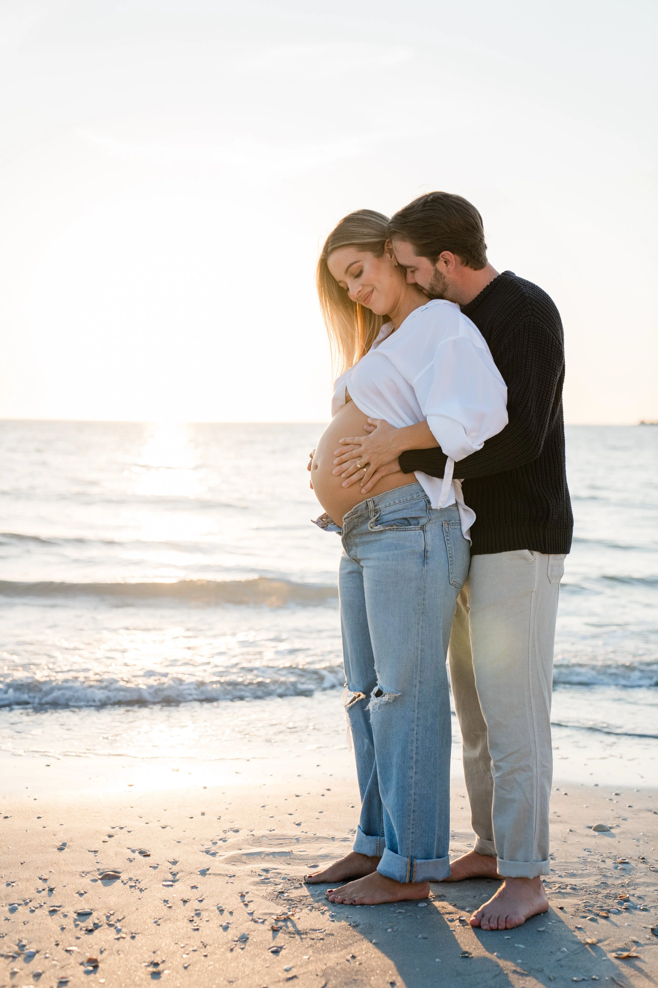 A pregnant woman and her partner standing barefoot on the beach at sunset, embracing and smiling with the ocean in the background.