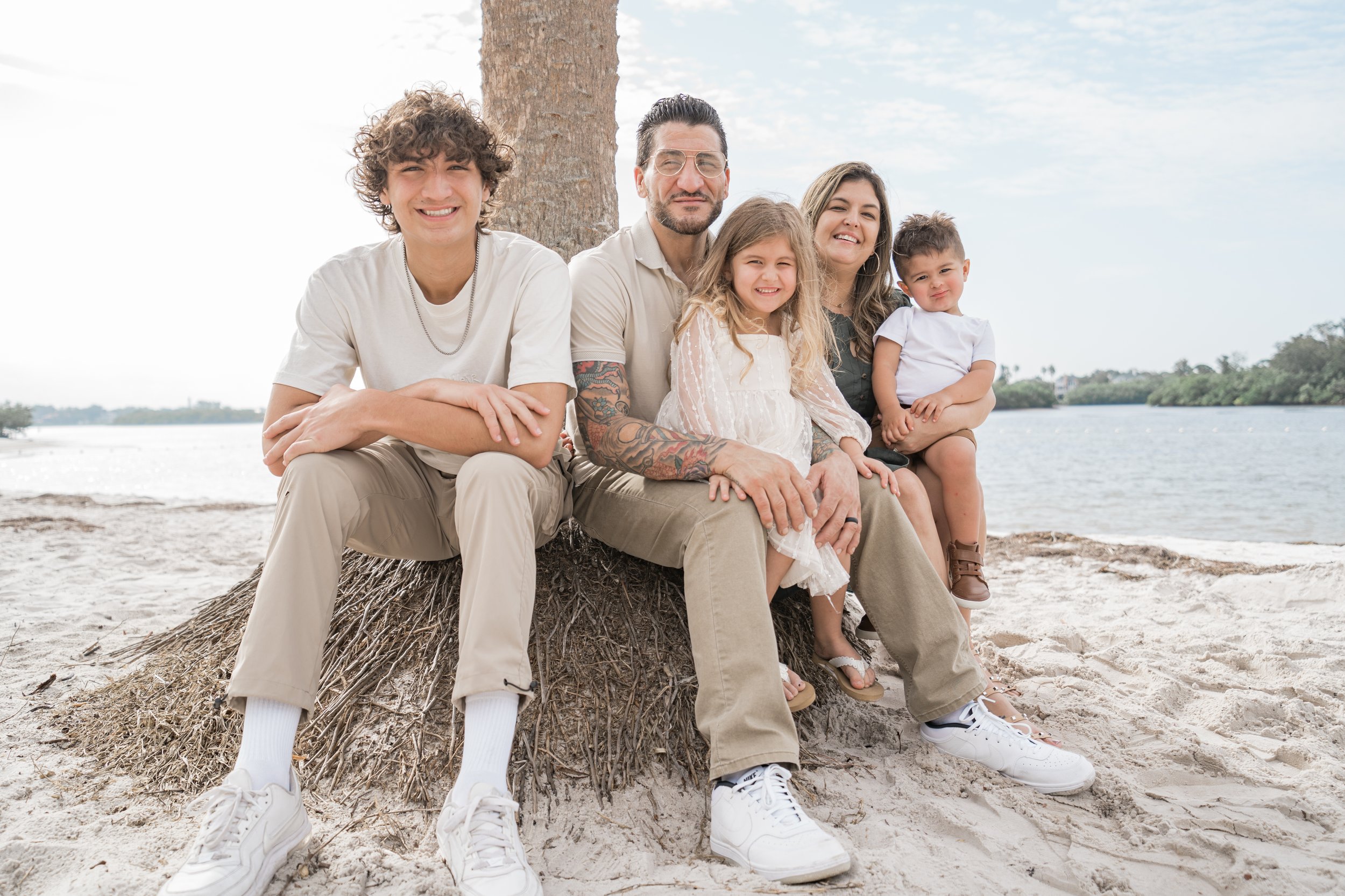 A family of six sitting on a tree root on a sandy beach near a river, smiling at the camera with a cloudy sky in the background.