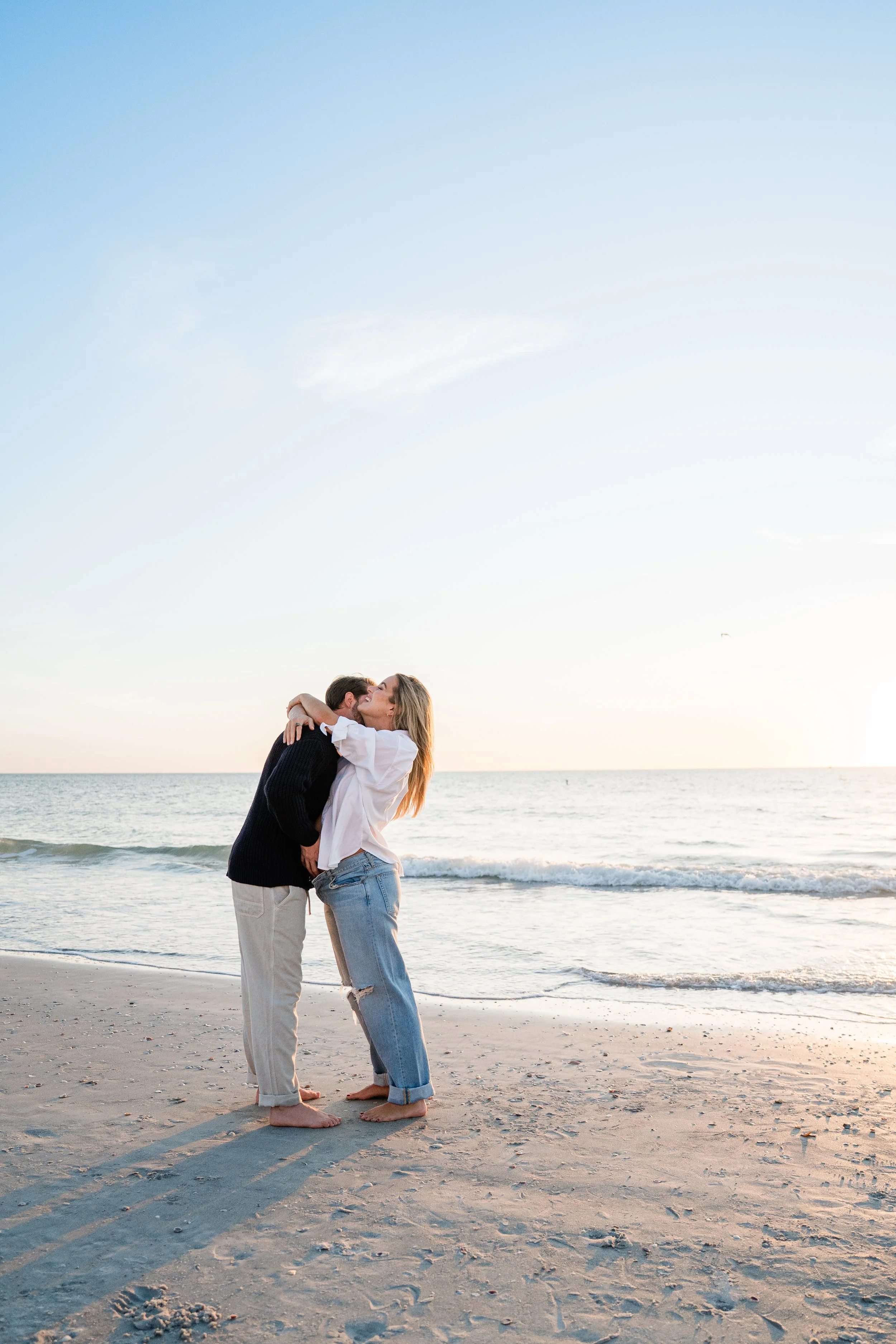 A couple kissing on the beach during sunset, with gentle waves and a clear sky.