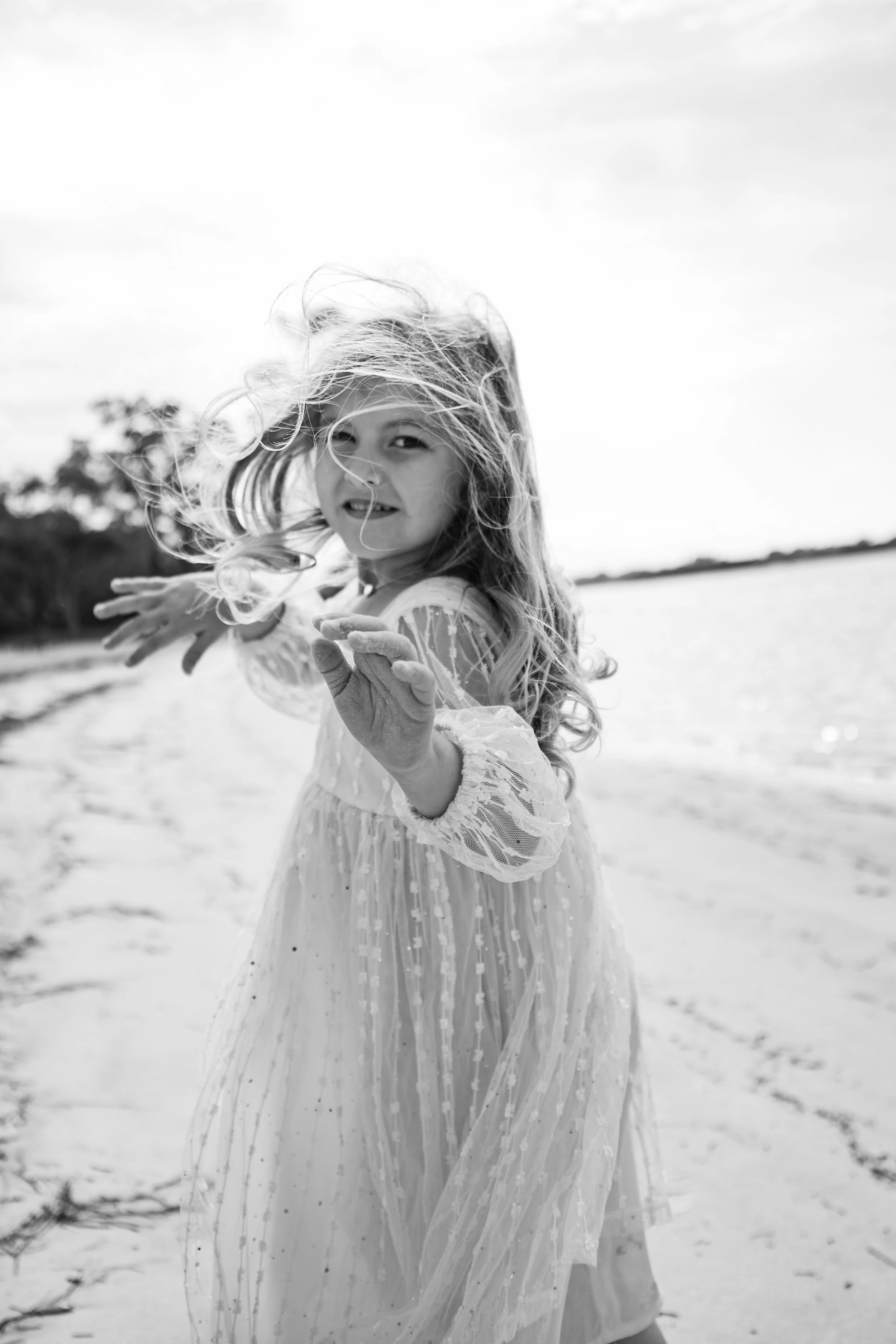 A young girl with flowing hair wearing a light, long dress, standing on a sandy beach with water in the background, smiling and playfully reaching out.