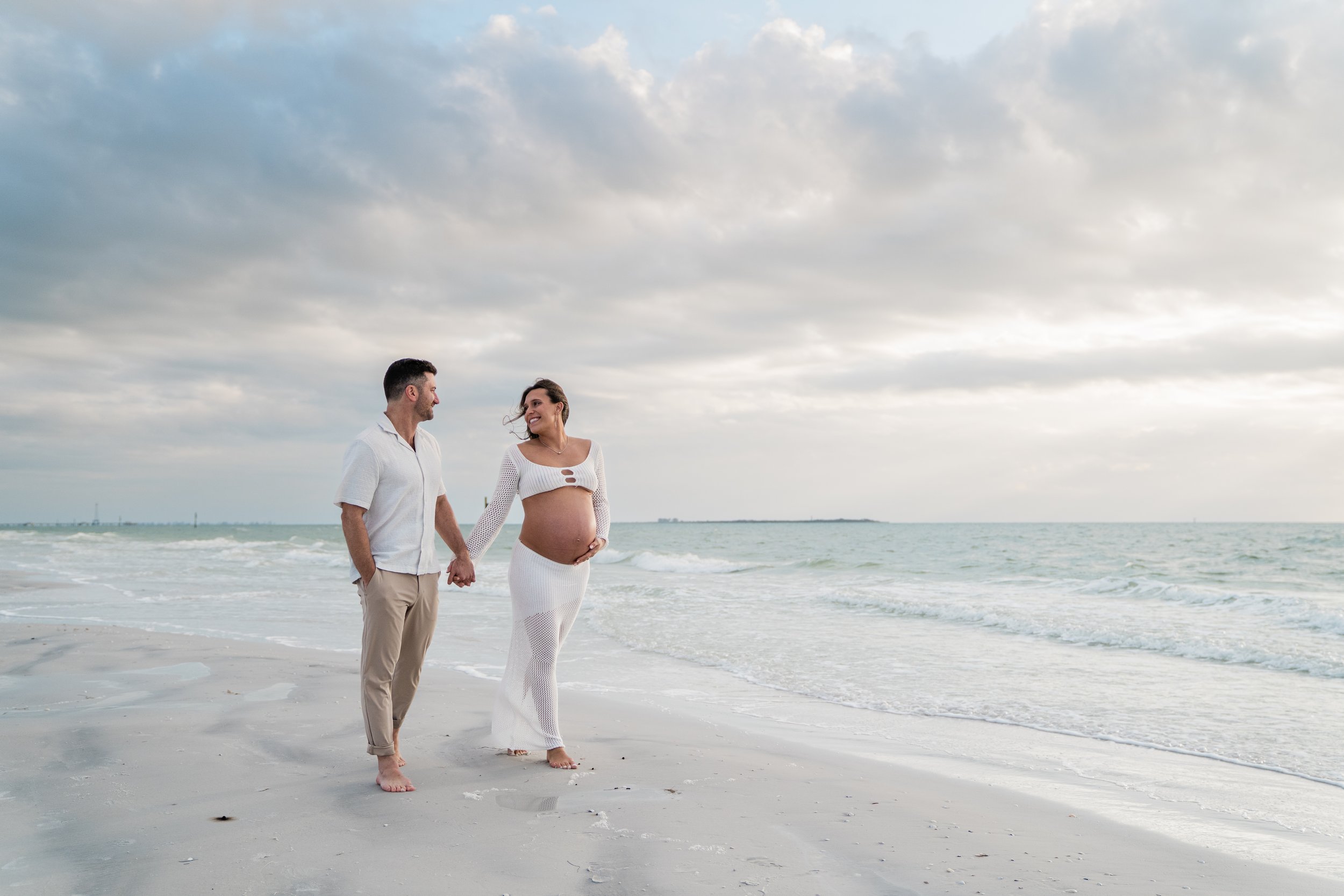 A pregnant woman and a man walking hand in hand along the beach at sunset, with the ocean and cloudy sky in the background.