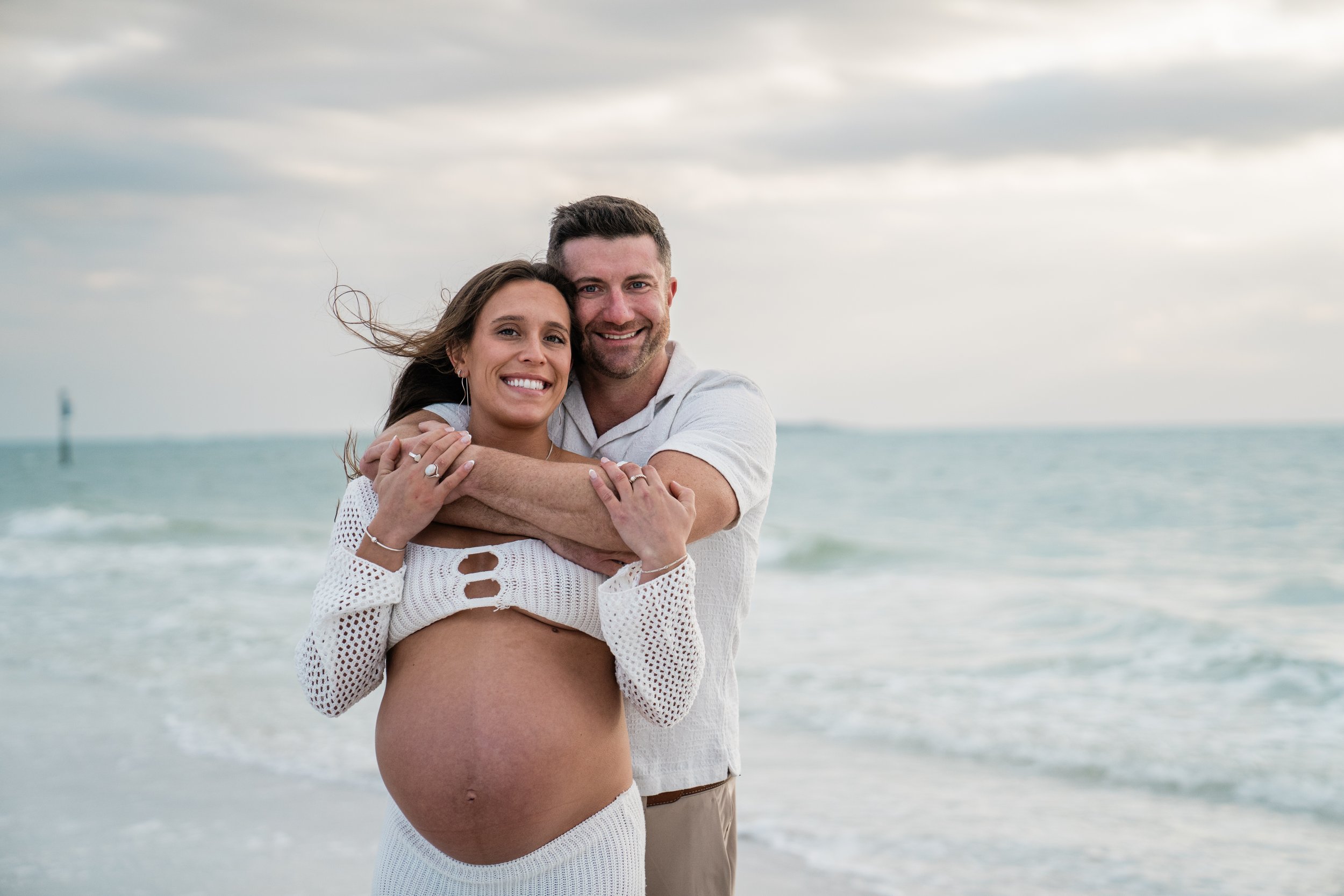 A happy couple, with the woman pregnant, standing on the beach near the ocean, embracing each other and smiling at the camera.