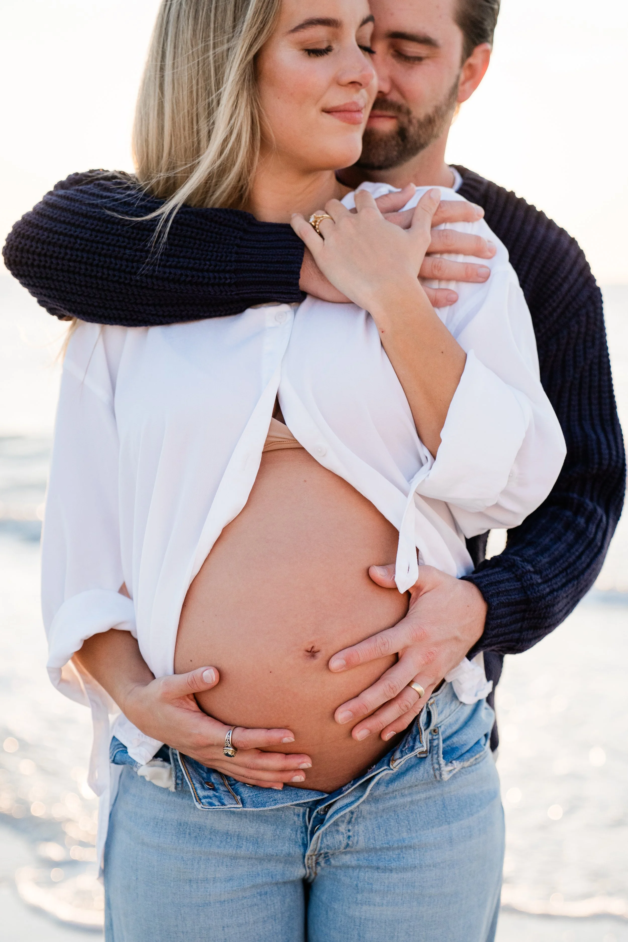 A pregnant woman with closed eyes, wearing an unbuttoned white shirt showing her belly, standing on a beach, embraced and kissed by a man from behind.