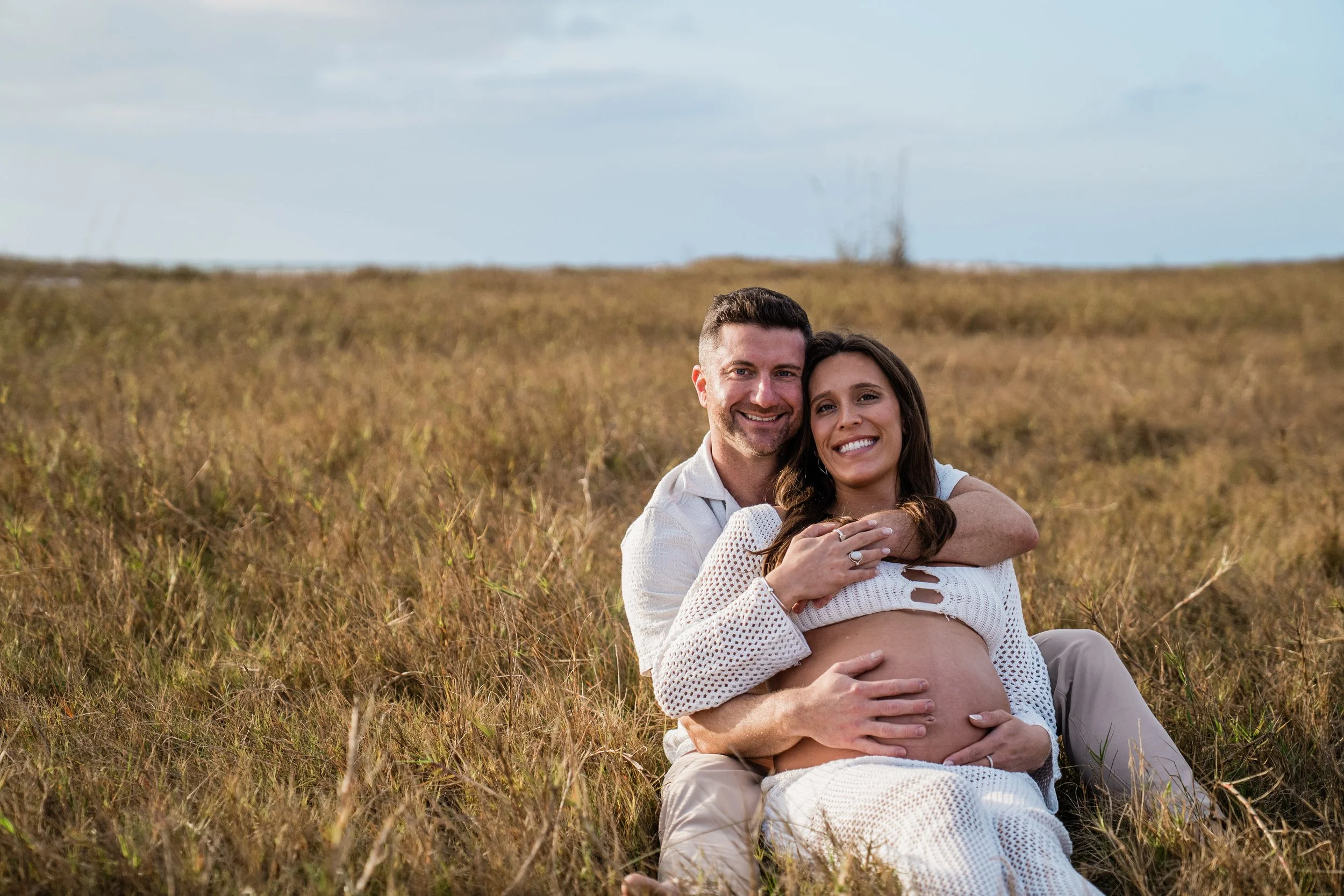 A happy pregnant woman sitting in a grassy field with her partner. They are both smiling and embracing, with the man's arms around the woman, and the woman's hands resting on her belly. The sky is partly cloudy.