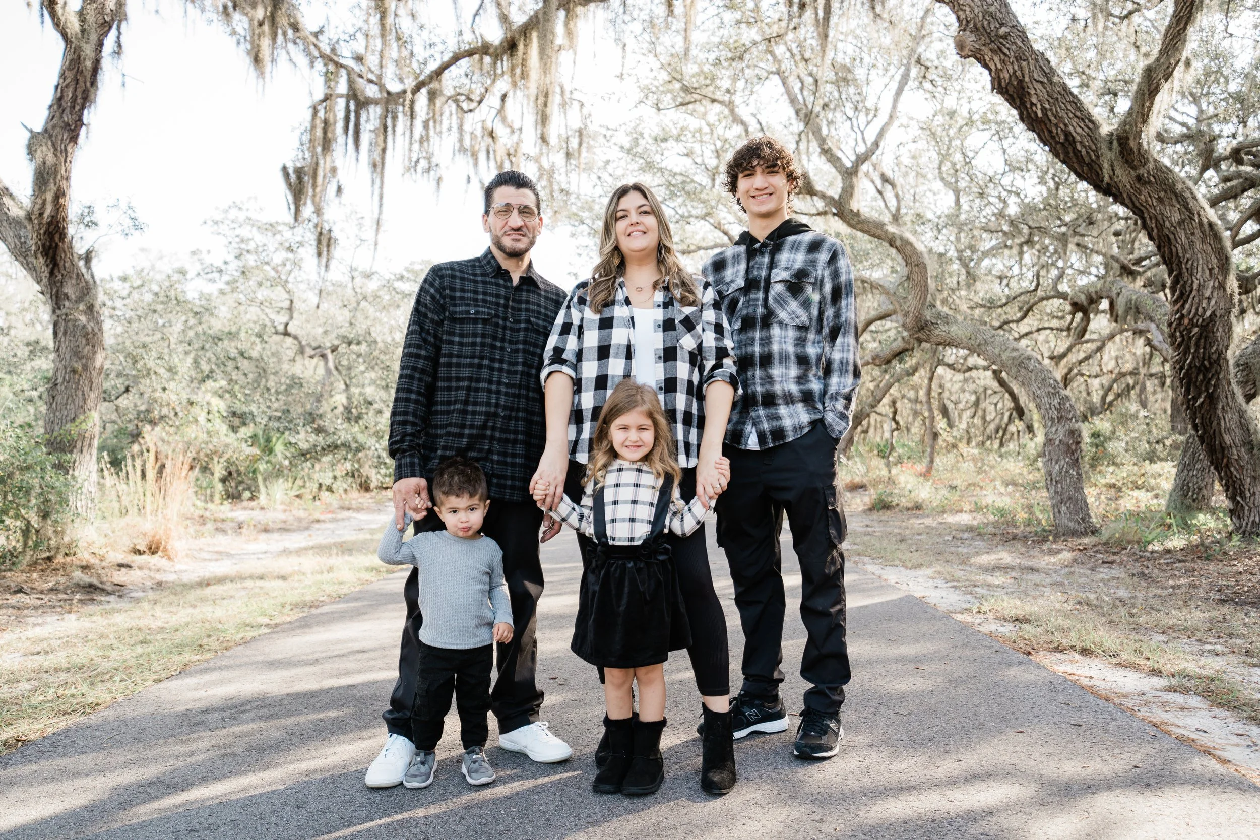 Family of five holding hands and walking on a paved path in a park surrounded by trees.