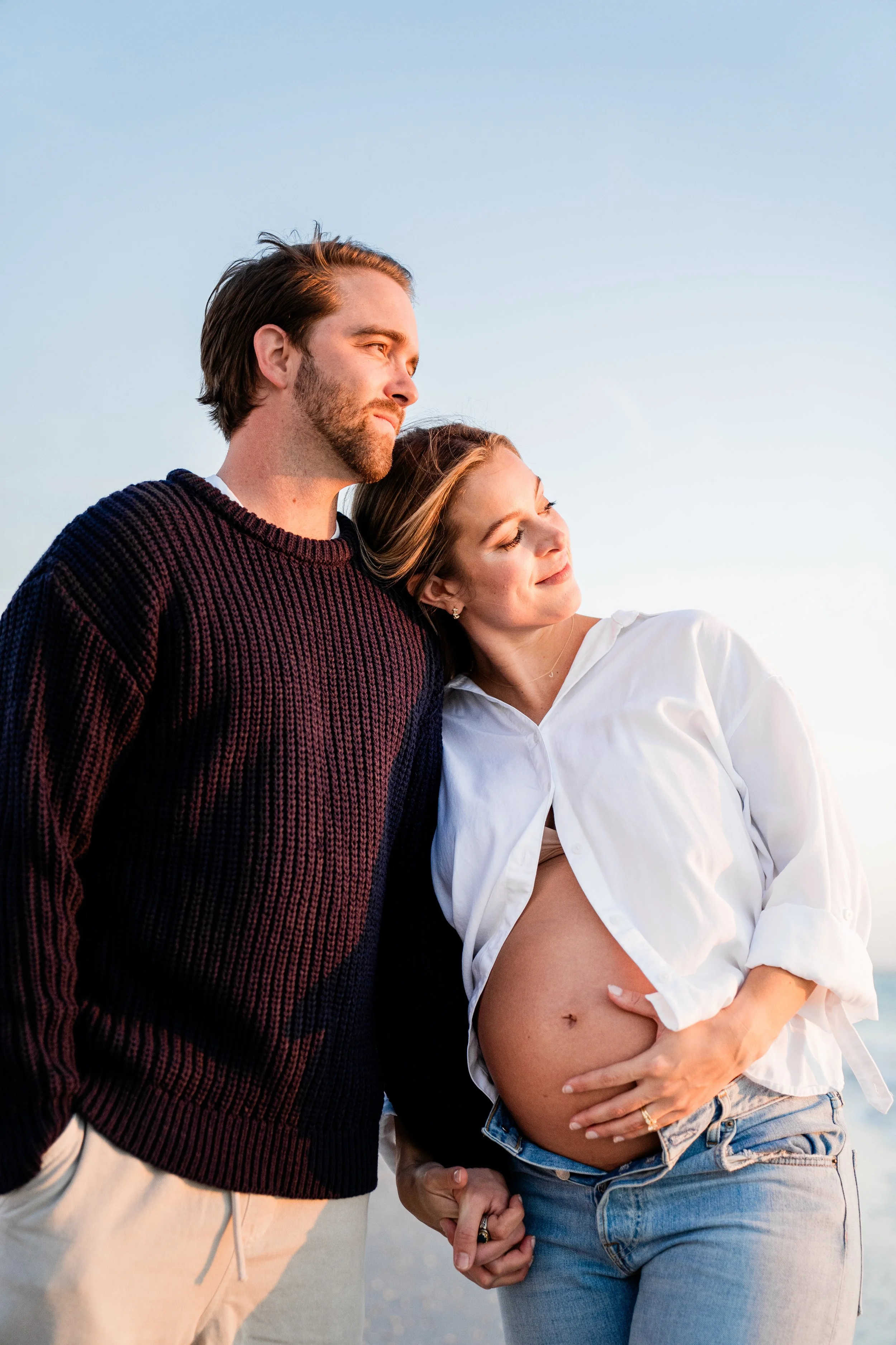 A pregnant woman and a man holding hands on a beach during sunset, with the woman resting her head on the man's shoulder.