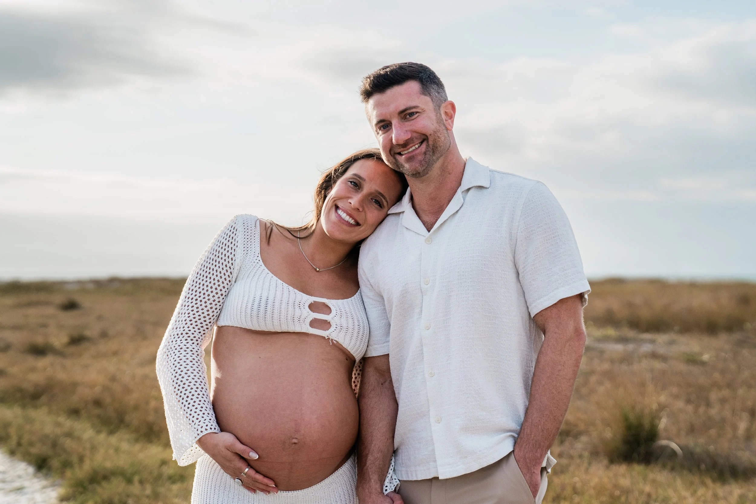 A pregnant woman and a man smiling and standing close together outdoors during daytime, with a cloudy sky in the background.