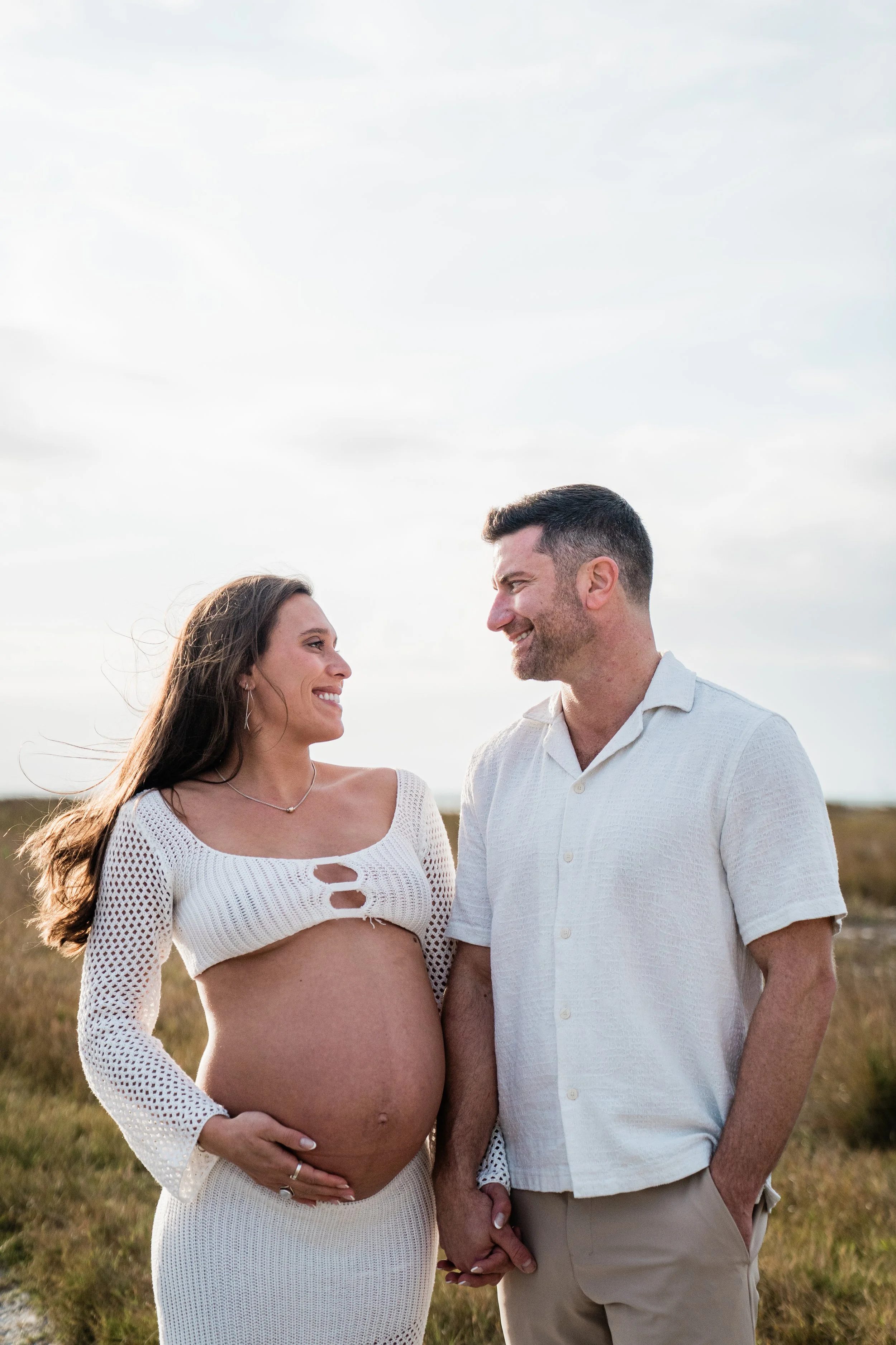 A pregnant woman and a man hold hands and look at each other outdoors against a cloudy sky.
