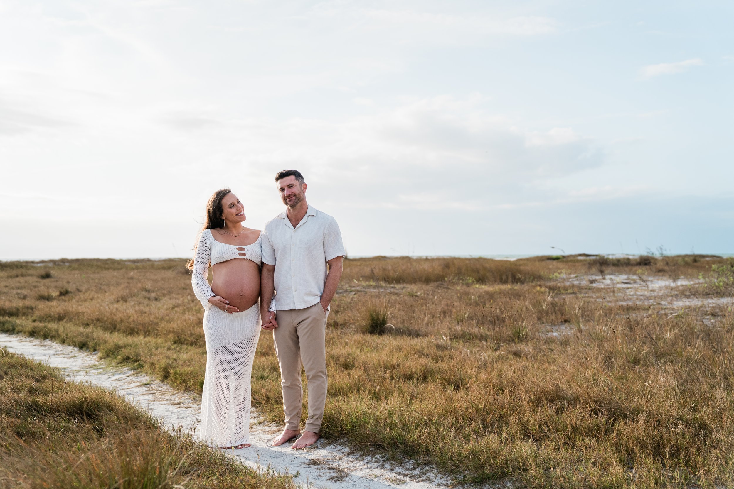 Pregnant woman and man standing hand in hand on a sandy path in a grassy field with a partly cloudy sky.