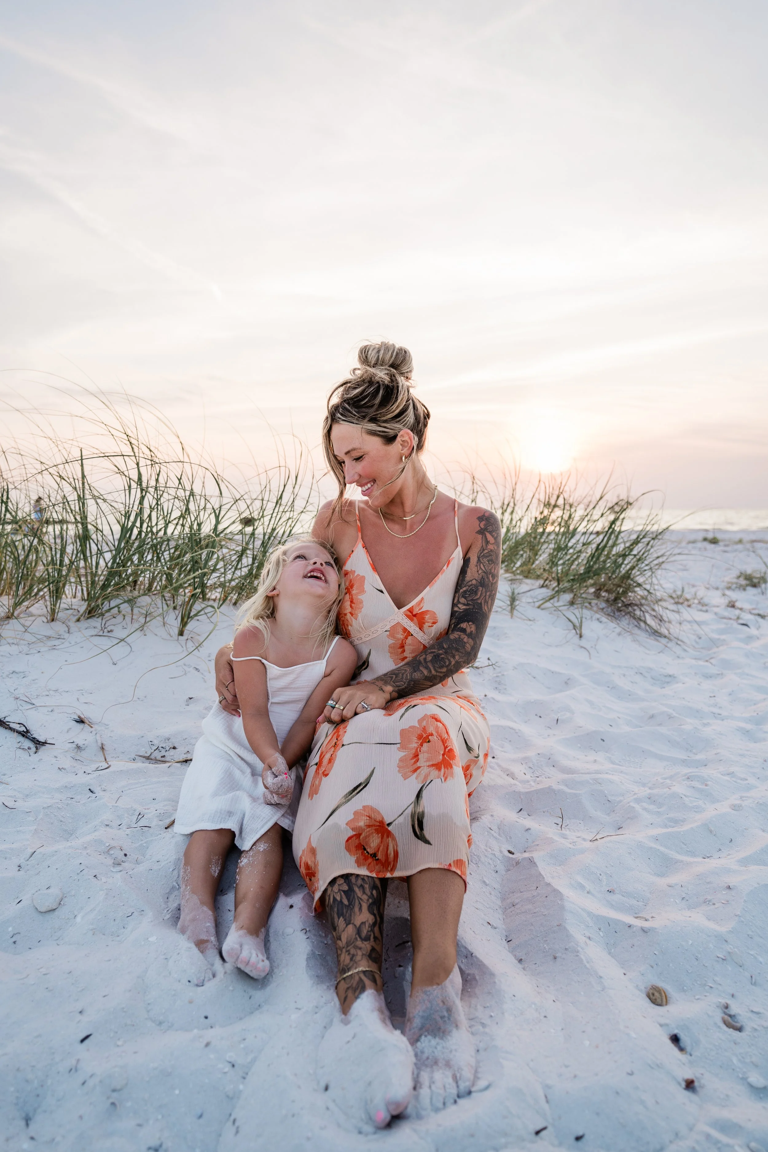 A woman and a young girl sitting on the sandy beach, smiling and looking at each other during sunset. The woman has tattoos on her arm and leg, and the girl is wearing a white dress. There are beach grass and a cloudy sky in the background.
