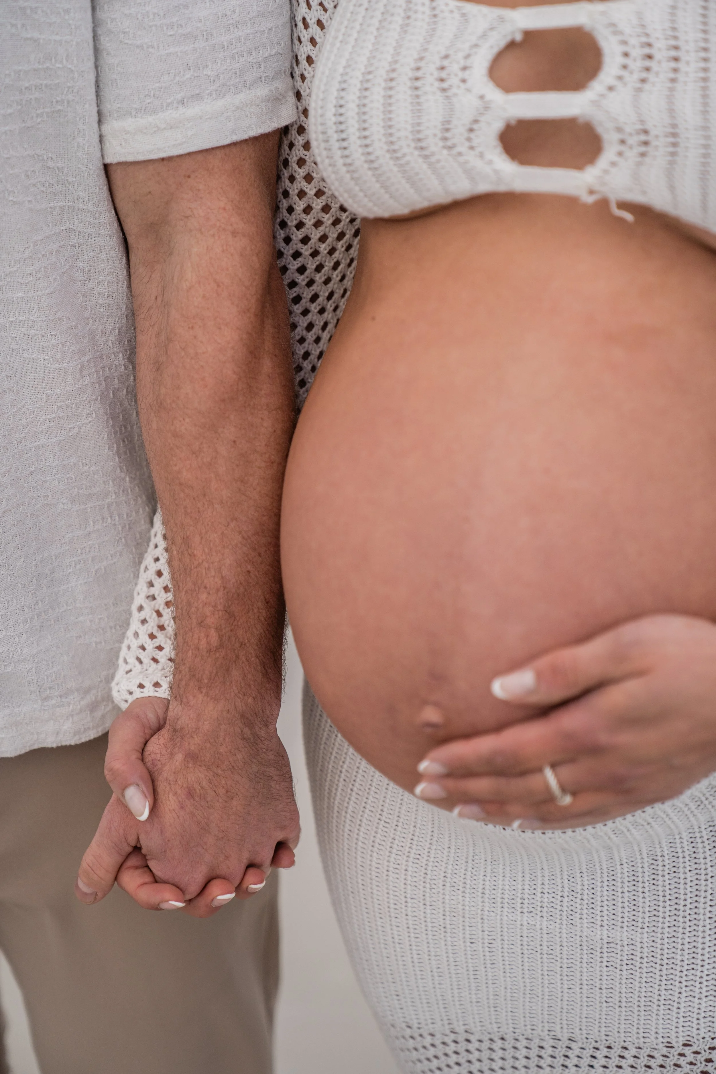 A woman with a pregnant belly being held by a man, both dressed in white. The woman is wearing a white knit top with holes and a white knit skirt, and the man is wearing a white shirt and beige pants. They are holding hands and standing close togethe