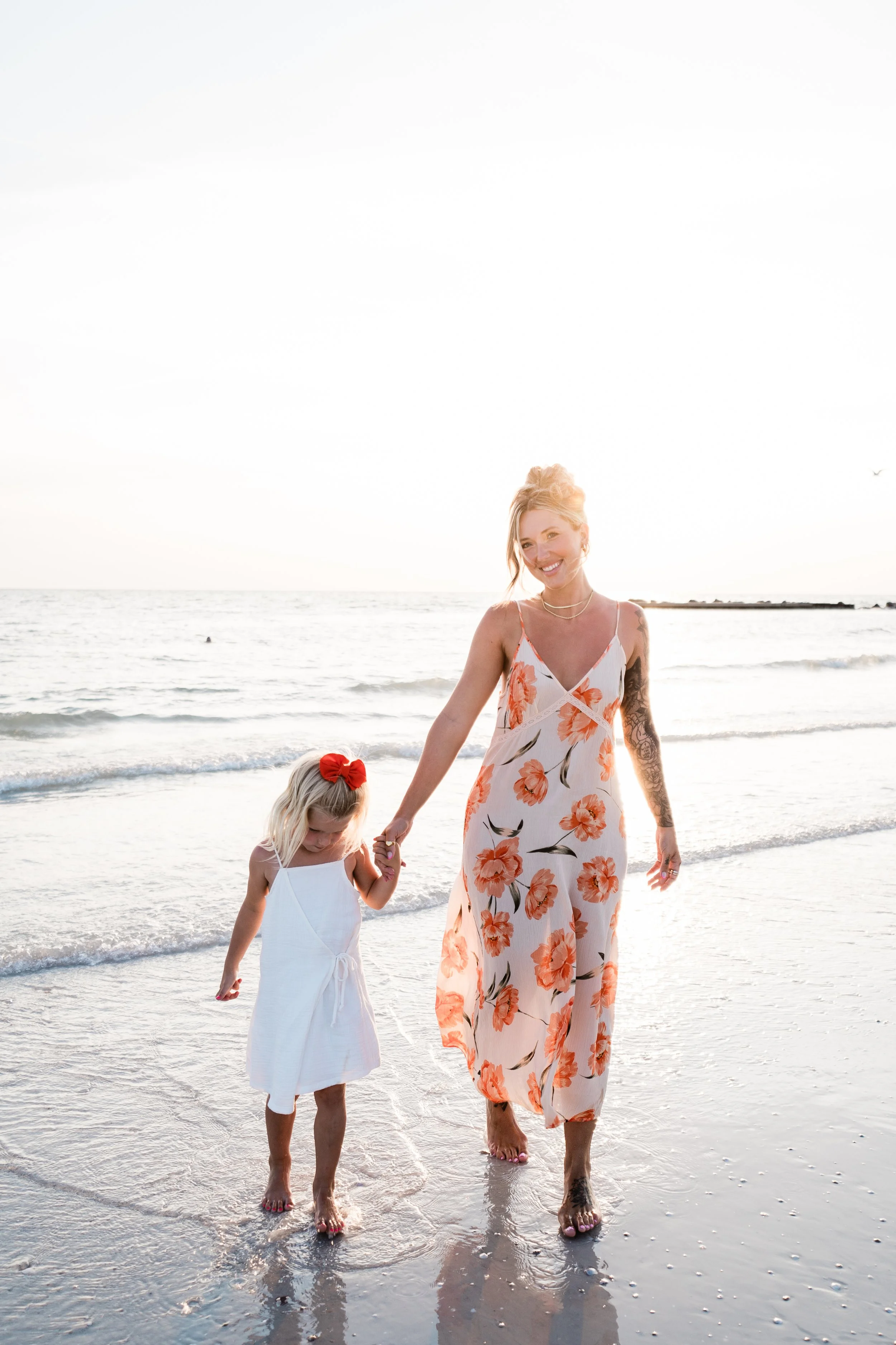 A woman and a young girl walking hand in hand along the beach at sunset, with waves in the background.