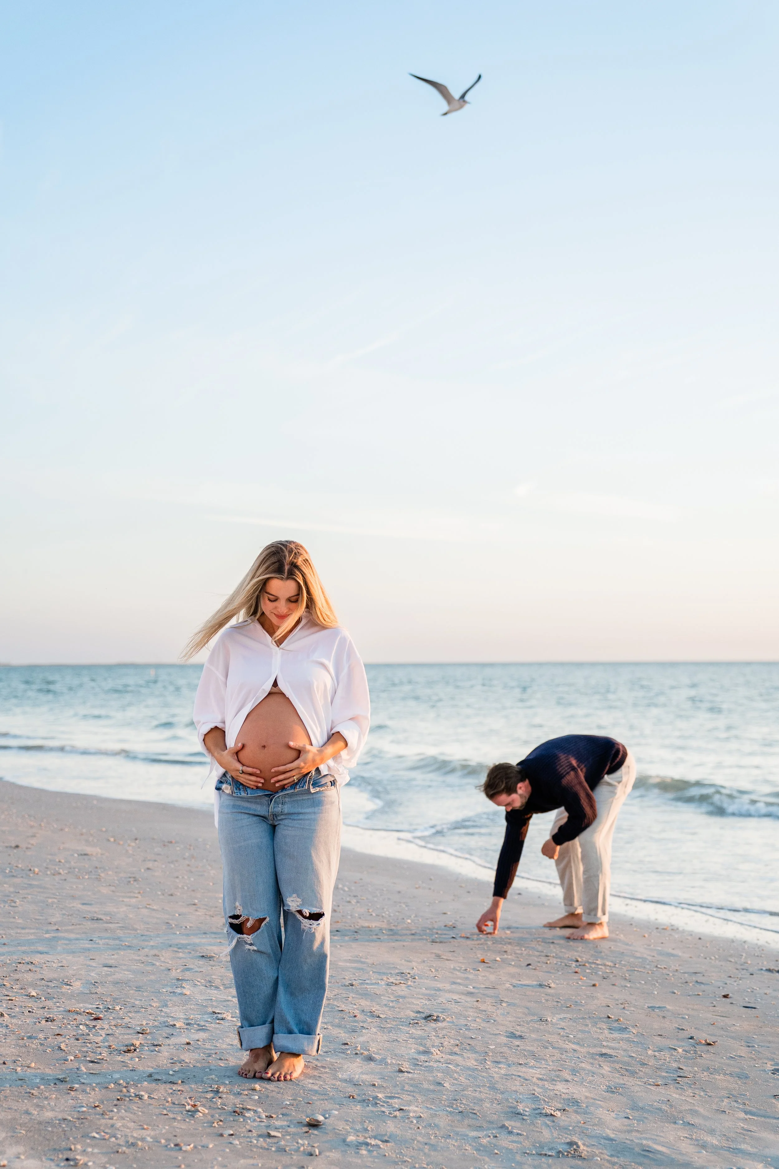 A pregnant woman in a white blouse and ripped jeans standing barefoot on a beach, smiling and gently holding her belly, with a man in a dark sweater and light pants crouching nearby, picking up seashells, during sunset with seagull flying in the sky.
