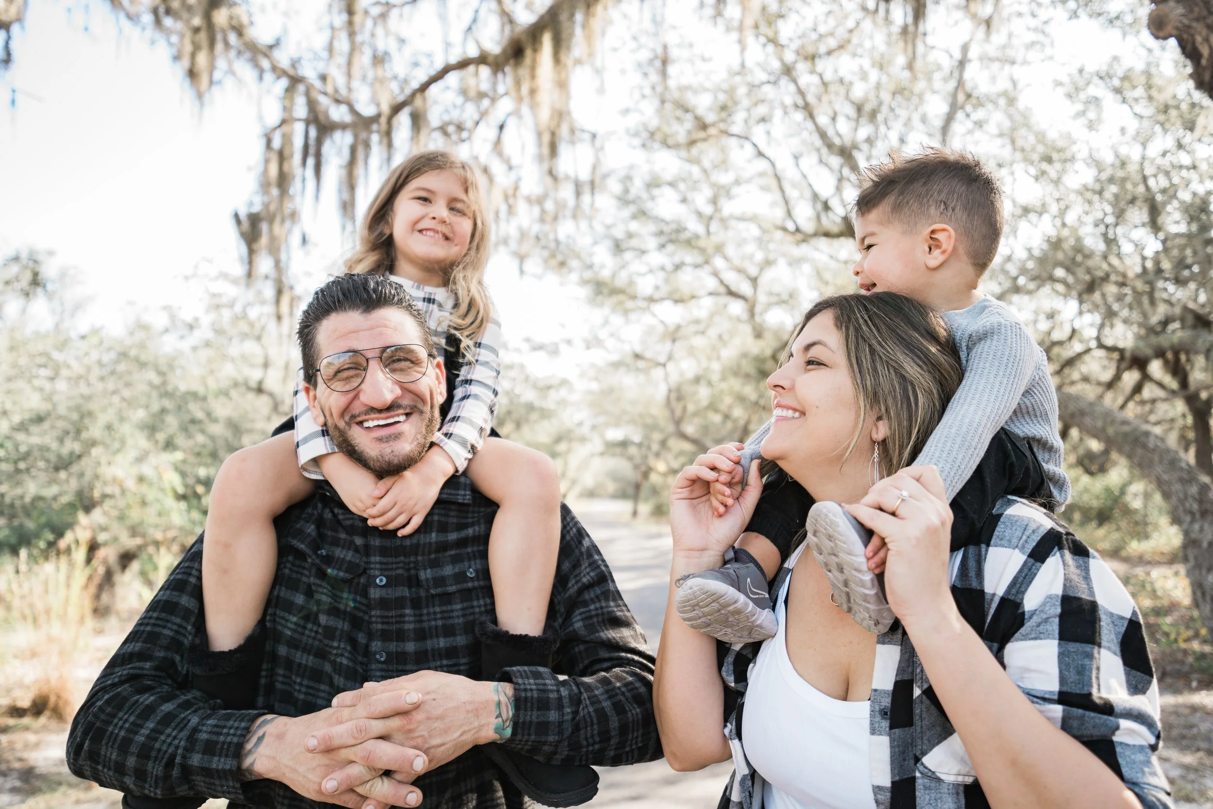 Family of four smiling outdoors with trees and Spanish moss in the background. The father is carrying a young girl on his shoulders, and the mother is holding a young boy on her shoulders, both children are smiling.