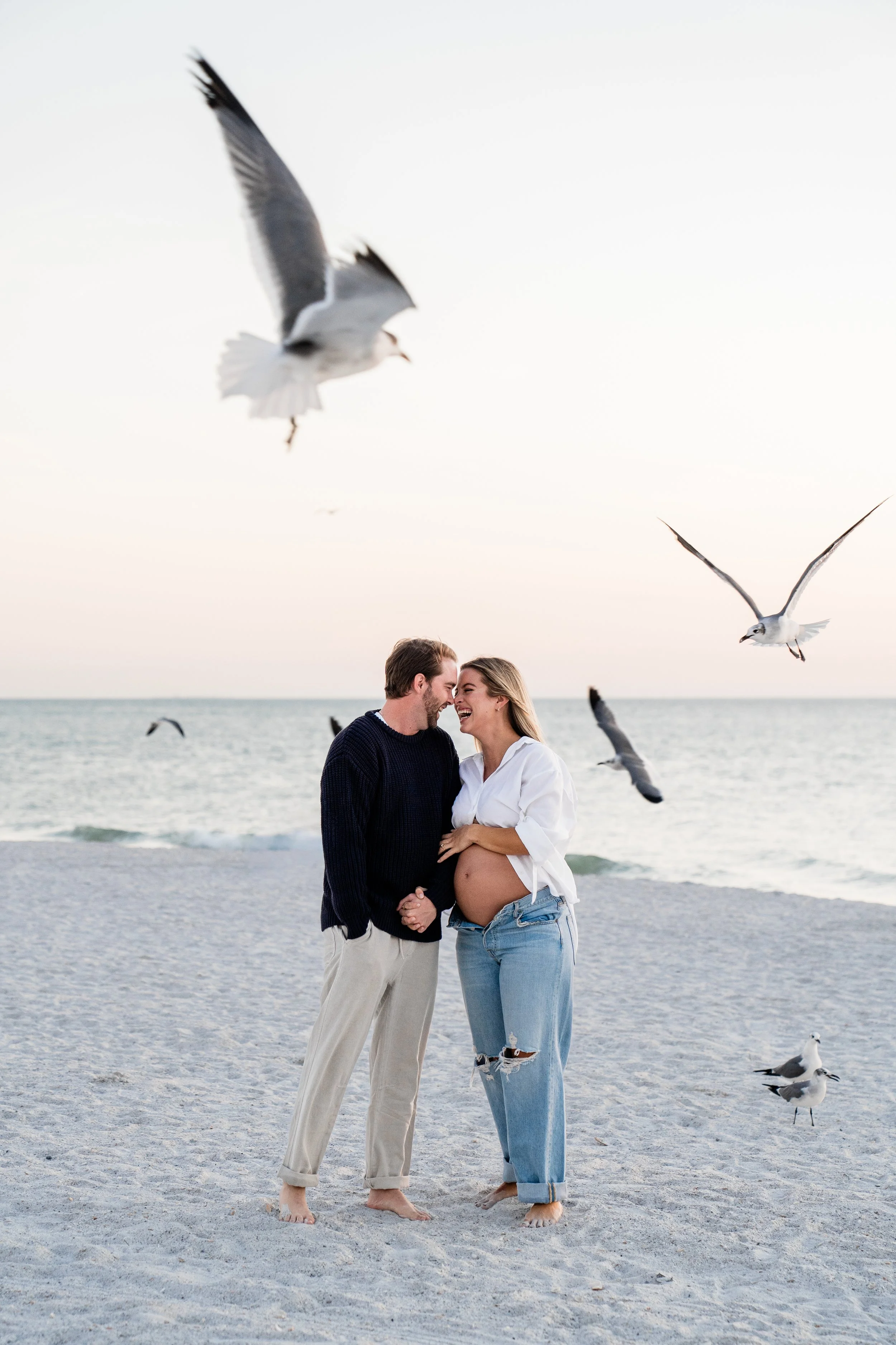 A pregnant woman and a man holding hands on a beach, surrounded by seagulls, with the ocean and sky in the background.