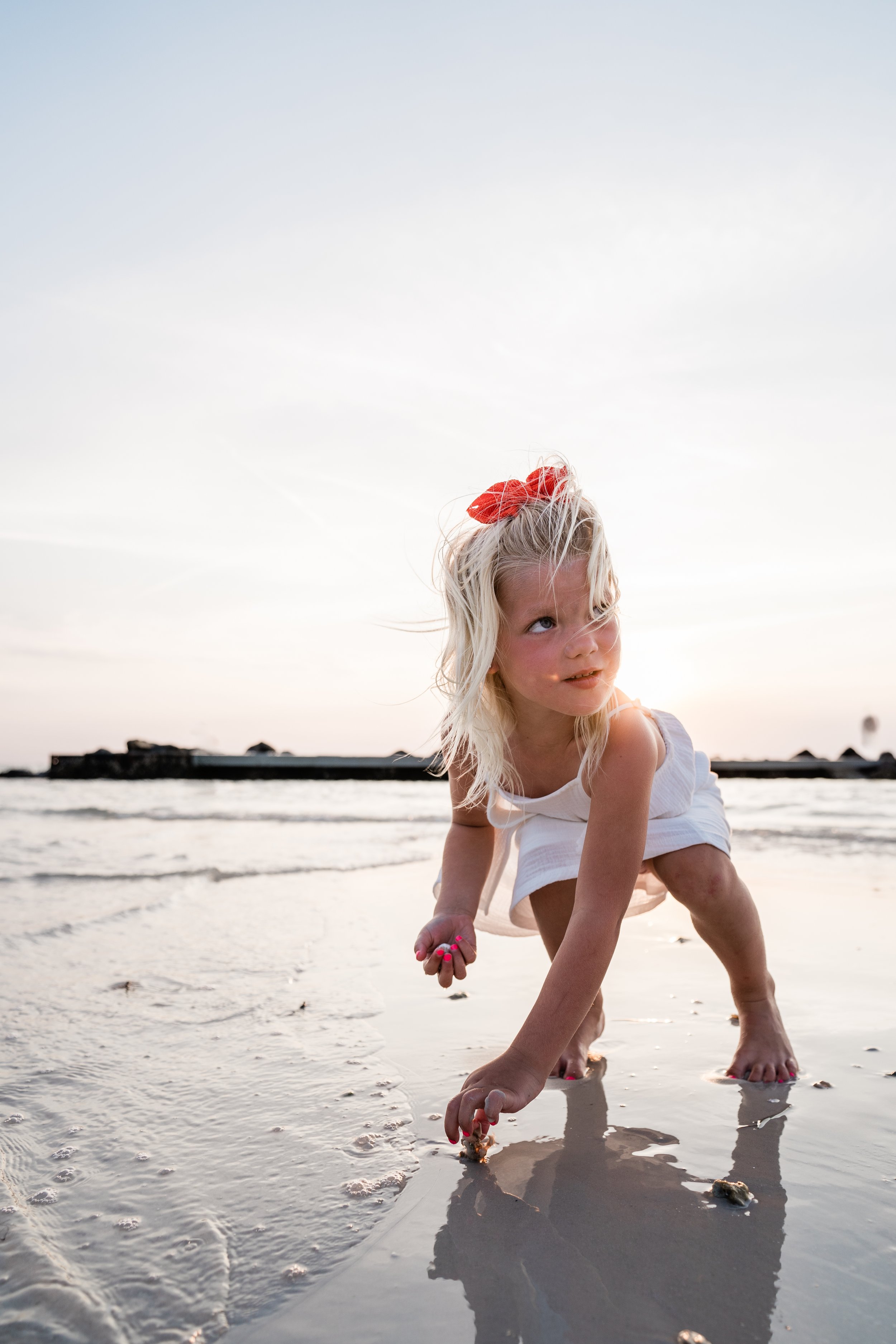 A young girl crouching in shallow water on a beach, with a red bow in her hair, wearing a white dress, during sunset.